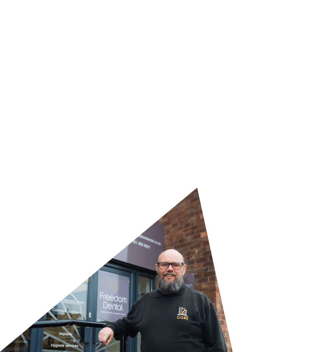 Smiling man with glasses and beard wearing a black CORE sweatshirt standing in front of Freedom Dental clinic entrance.