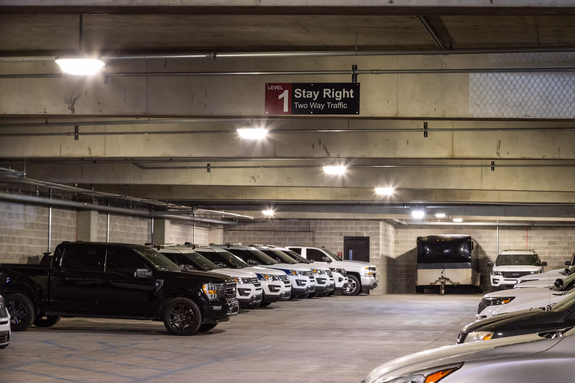 Indoor parking lot with multiple trucks and cars parked under concrete ceiling lights.
