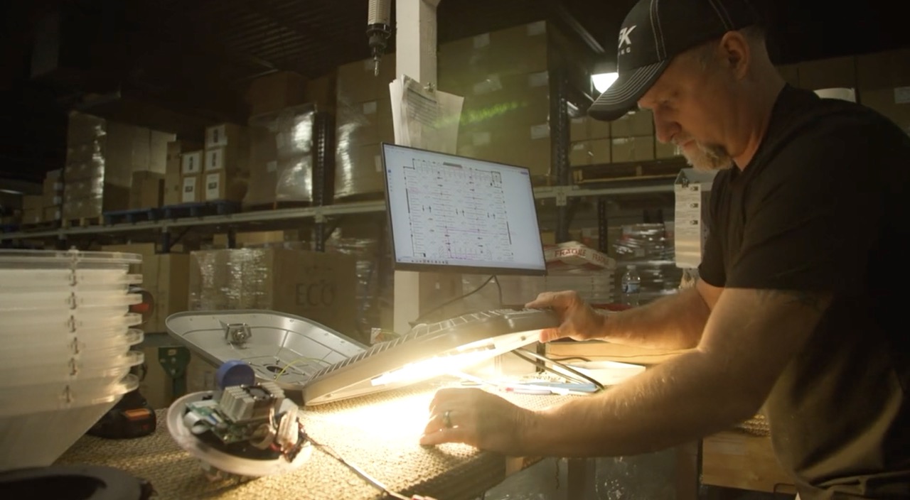Man inspecting and assembling an illuminated electronic device with a computer display in a warehouse.