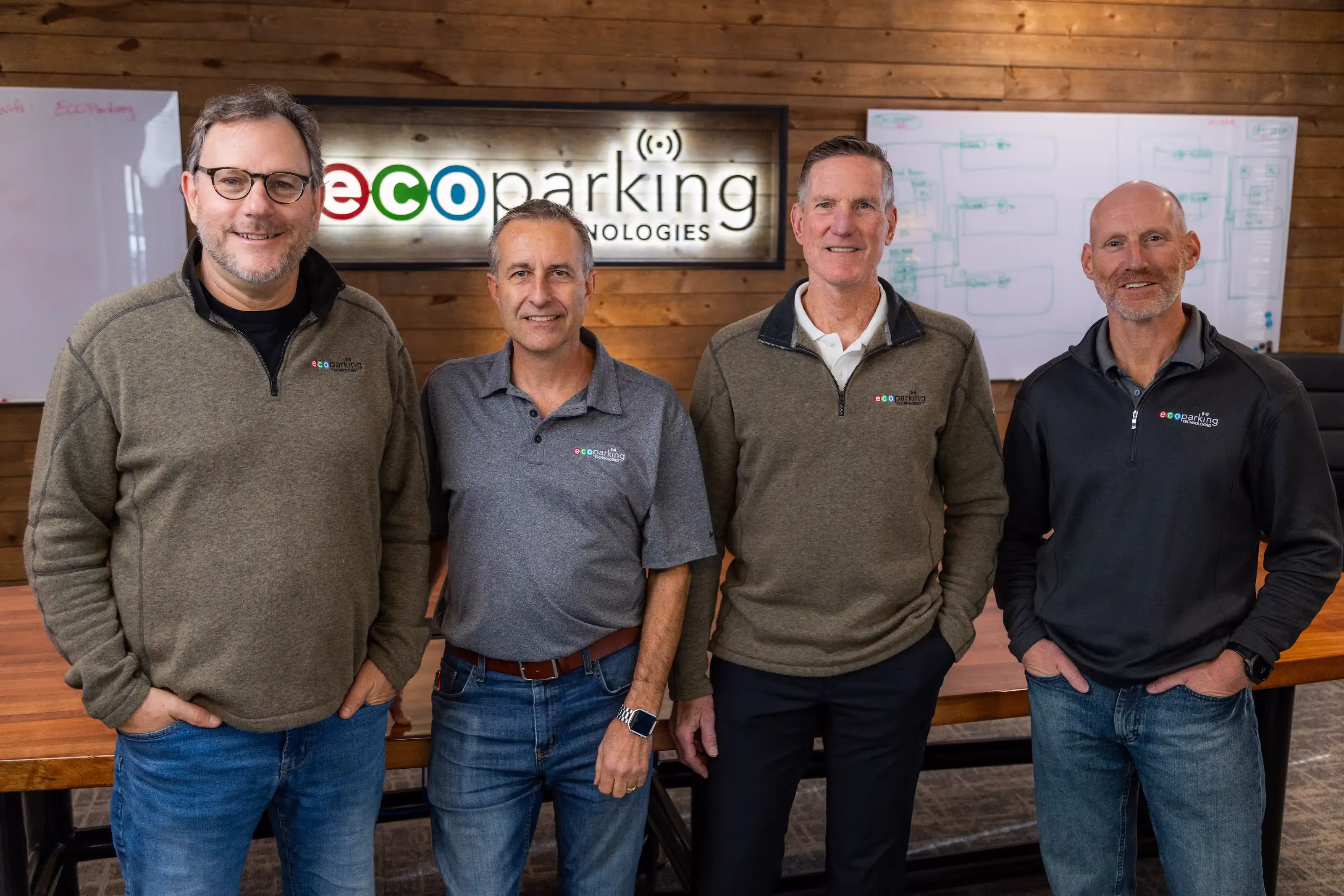Four men smiling in casual ecoParking Technologies branded tops standing in office in front of logo sign.