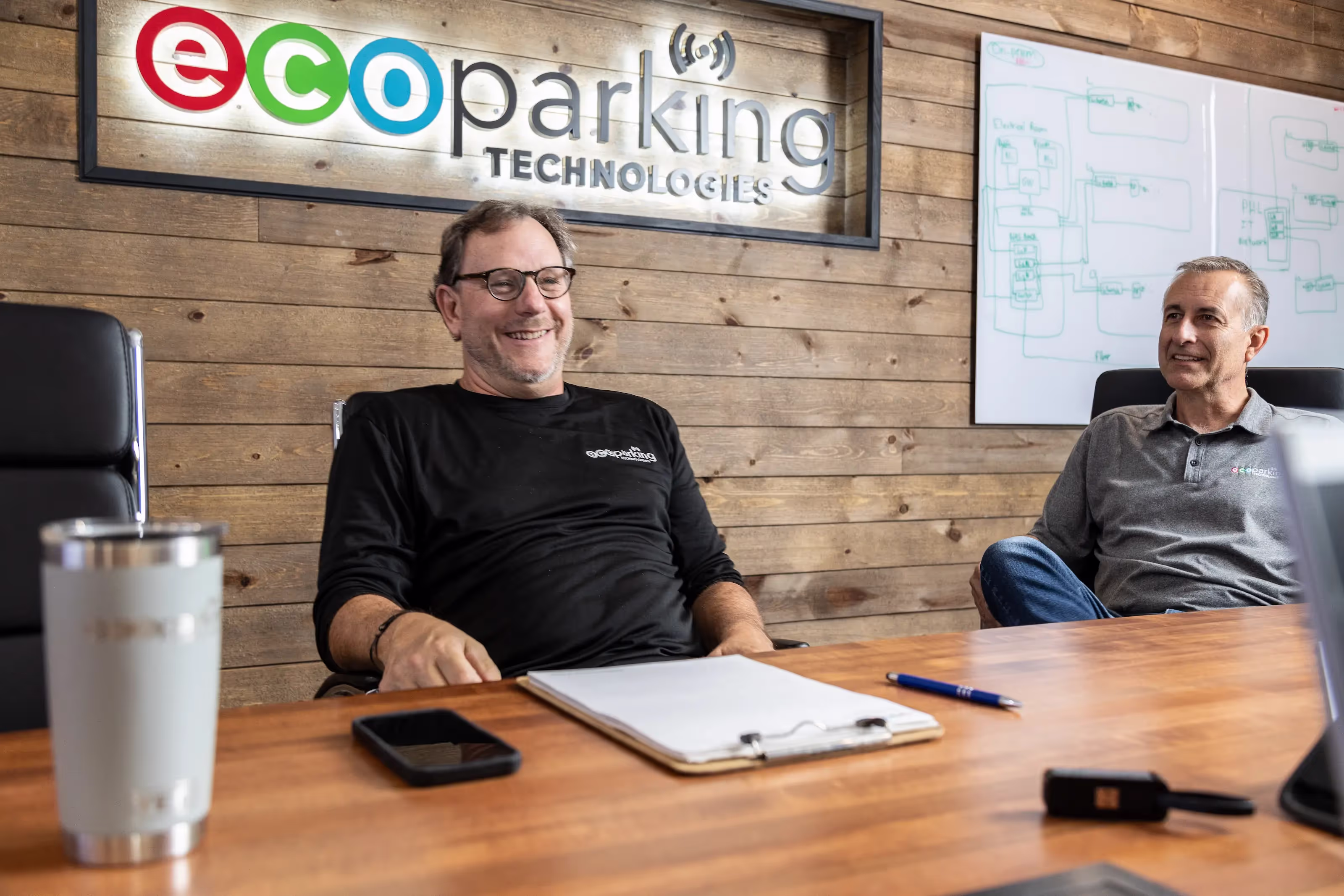 Two men seated at a wooden table in a meeting room with ecoparking logo on wall.