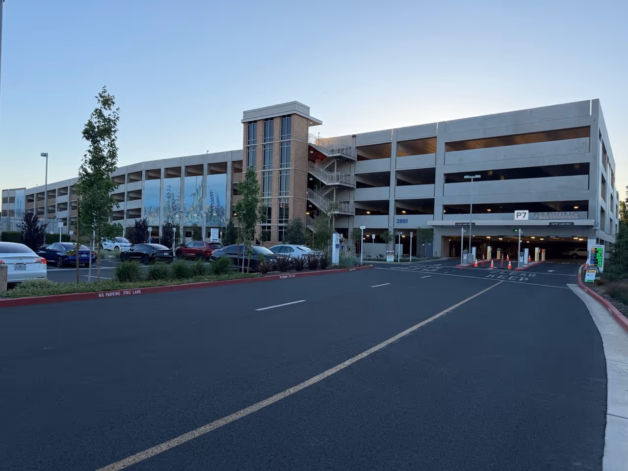 Multi-level parking garage with cars parked outside and an entrance with orange traffic cones.