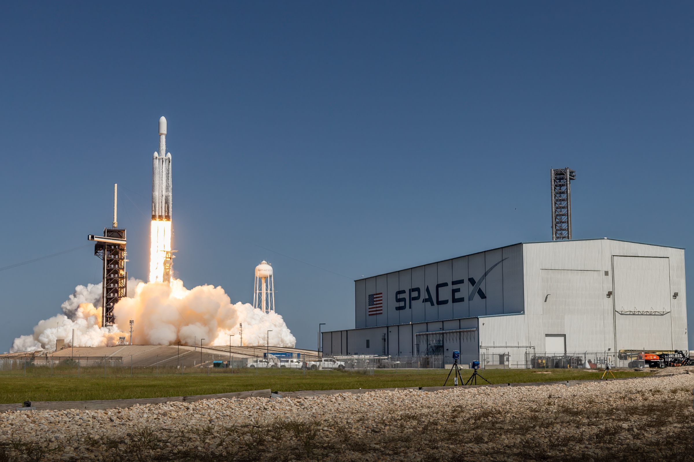 A Falcon Heavy lifts off from Launch Complex 39A with all three cores burning, seen past the SpaceX hangar.