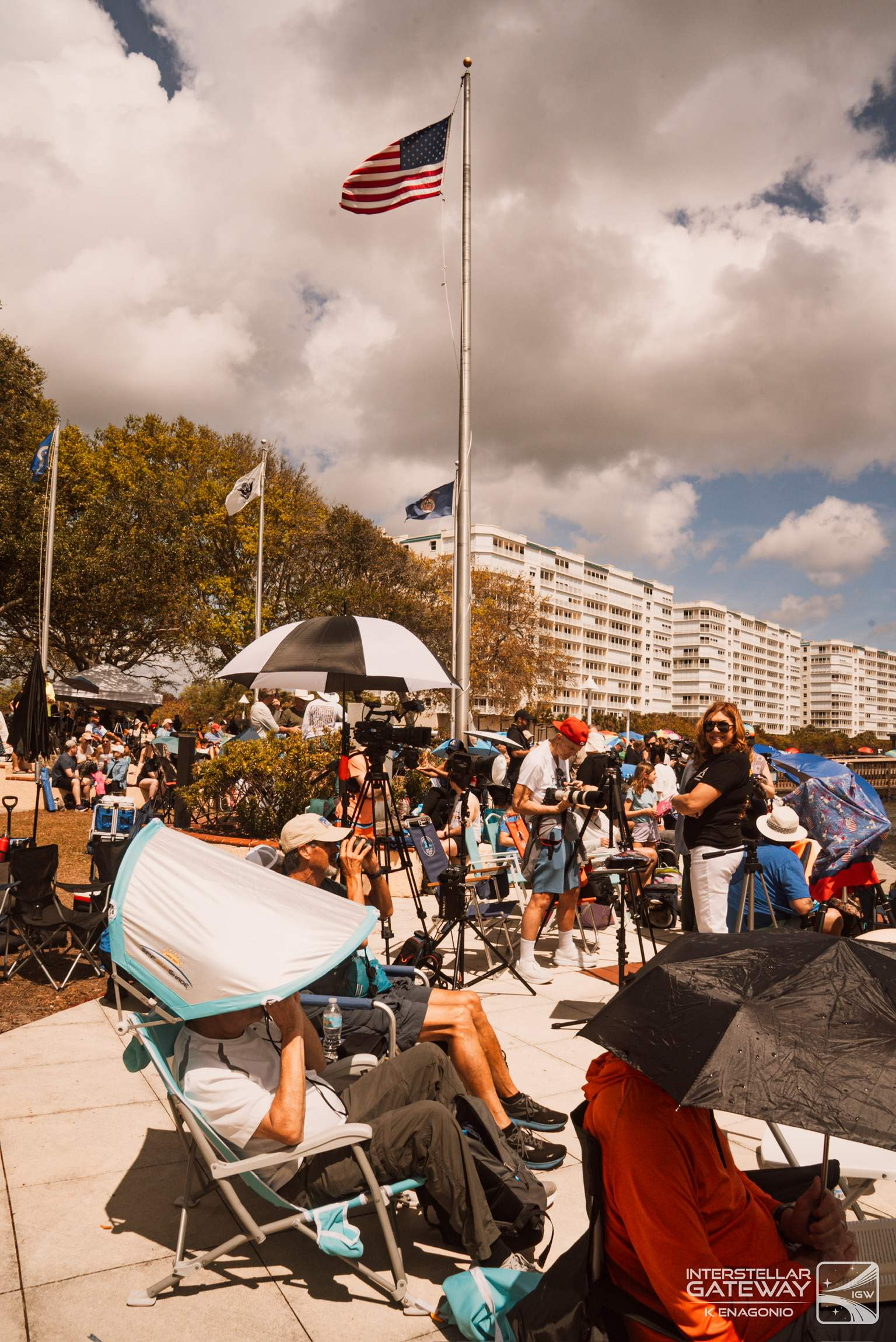 Spectators under umbrellas at a waterfront viewing site, a United States flag flying above them.