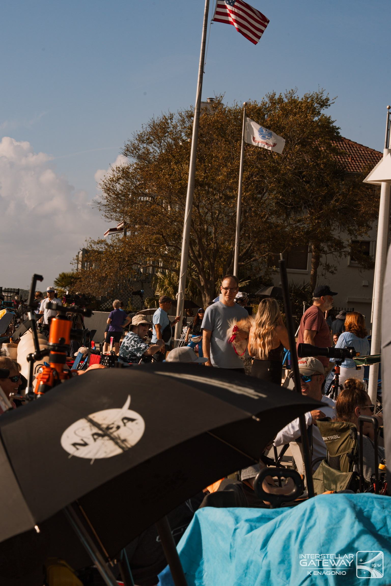 Spectators along a waterfront under flags, umbrellas up, waiting for a launch.
