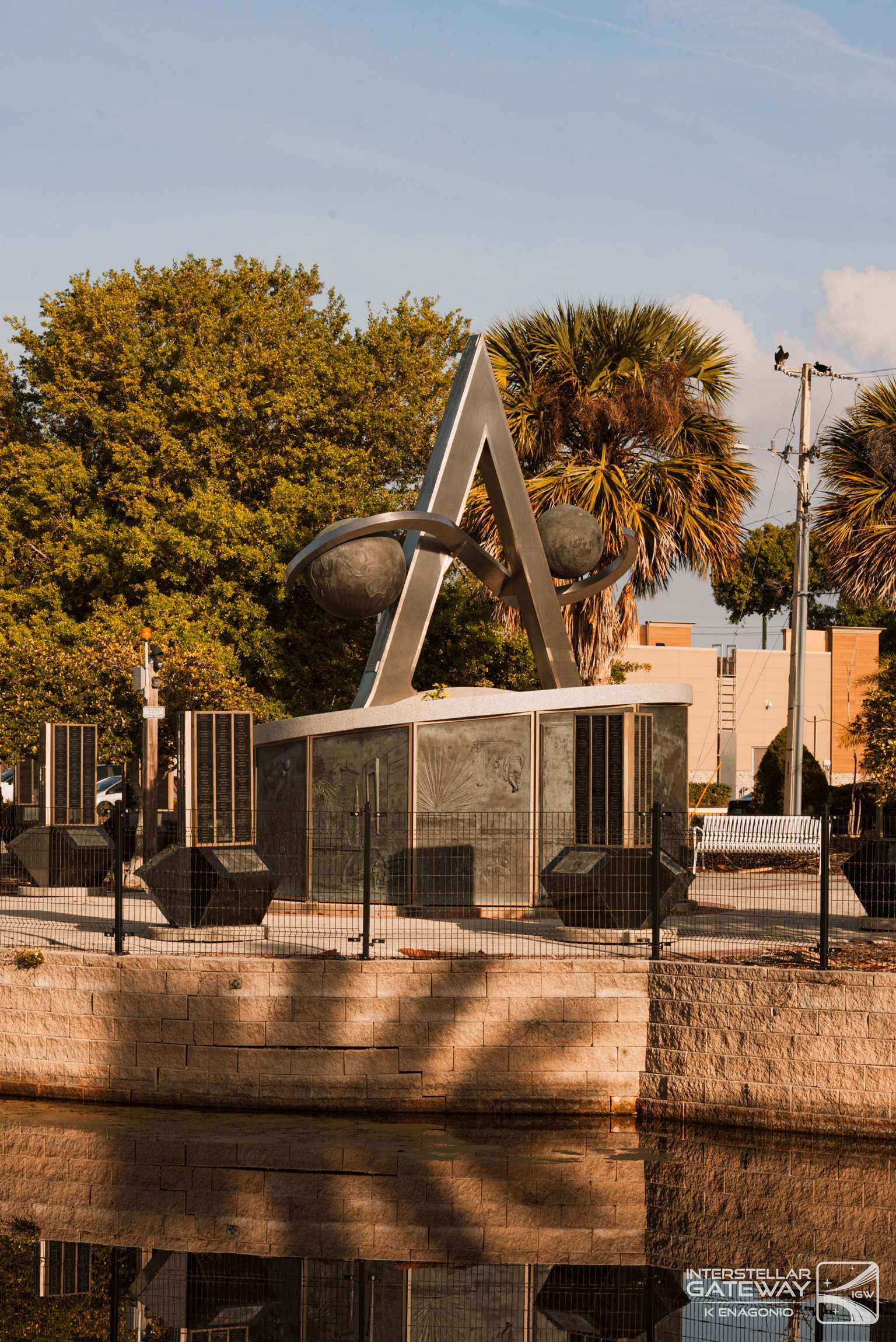 A stone monument with a stainless sculpture beside the water, palms behind it.