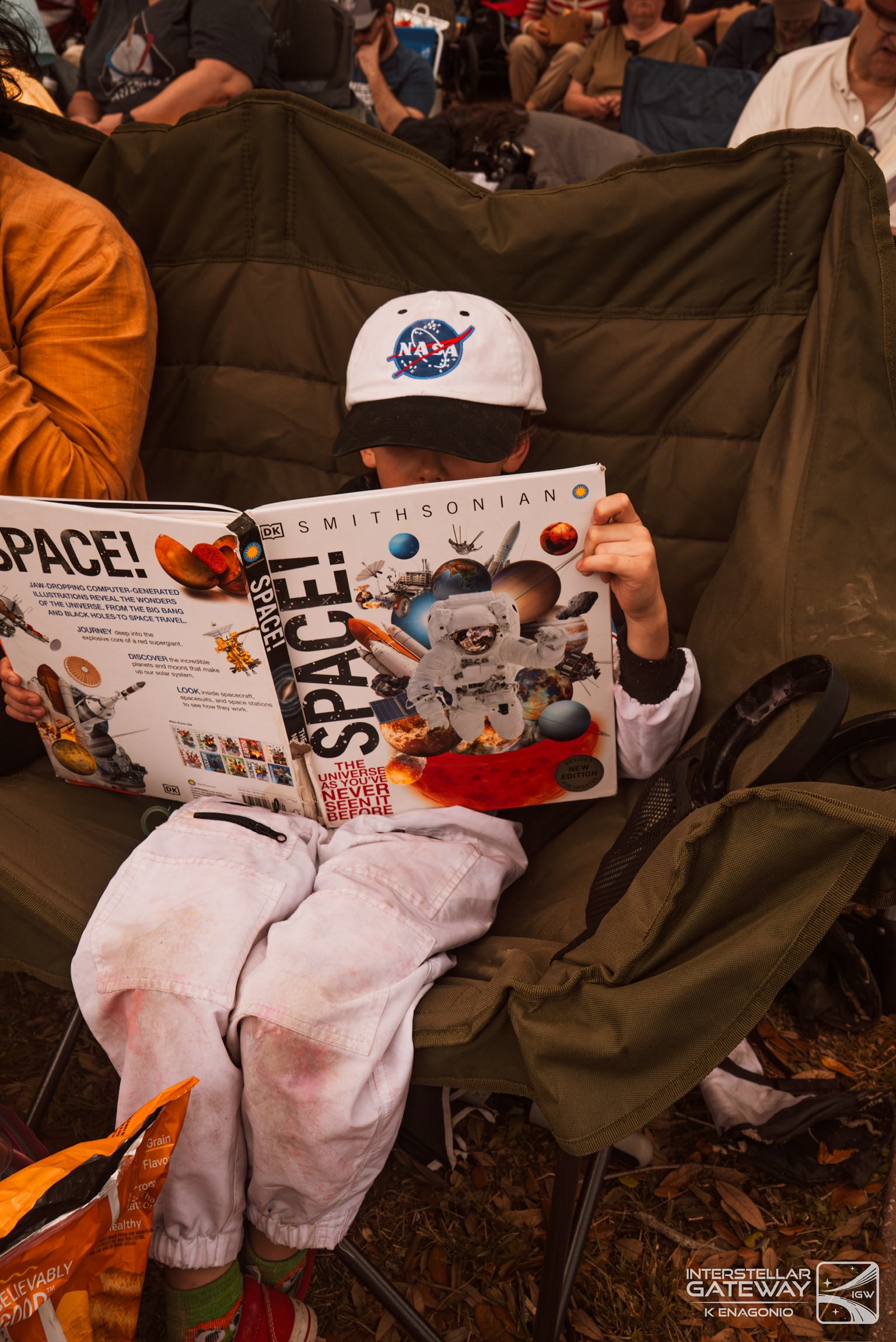 A child in a NASA cap reads a space magazine in a camp chair at a launch viewing site.