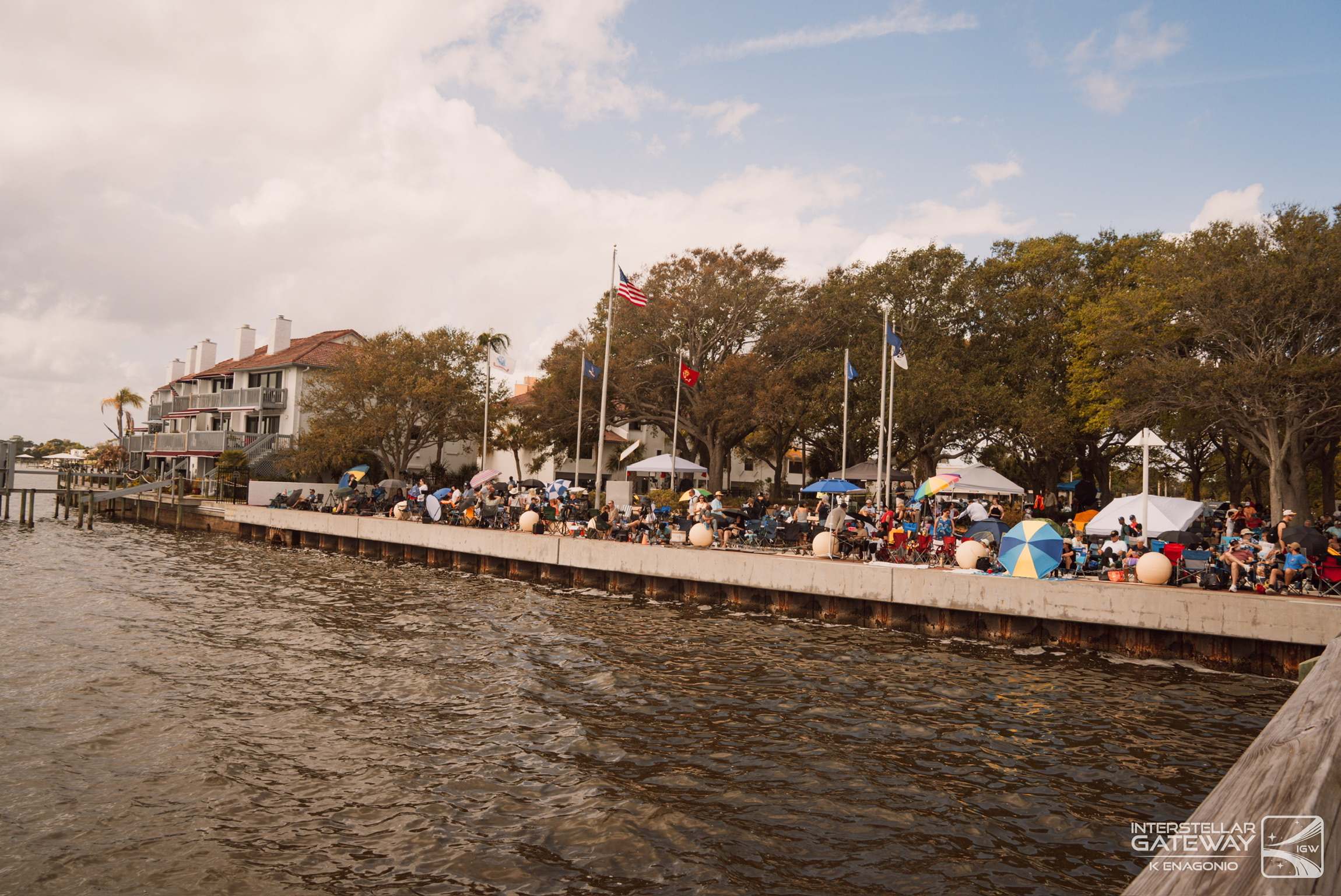 A crowd lines a waterfront under trees and flags, waiting for a launch.
