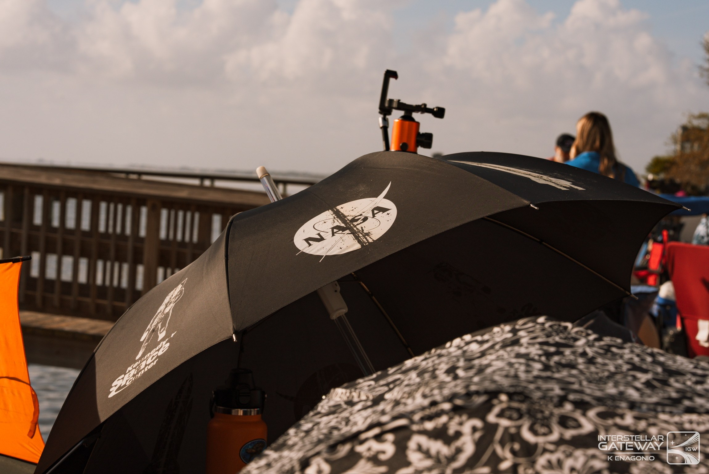 A NASA umbrella in the foreground of a crowded pier, cameras set up behind it.