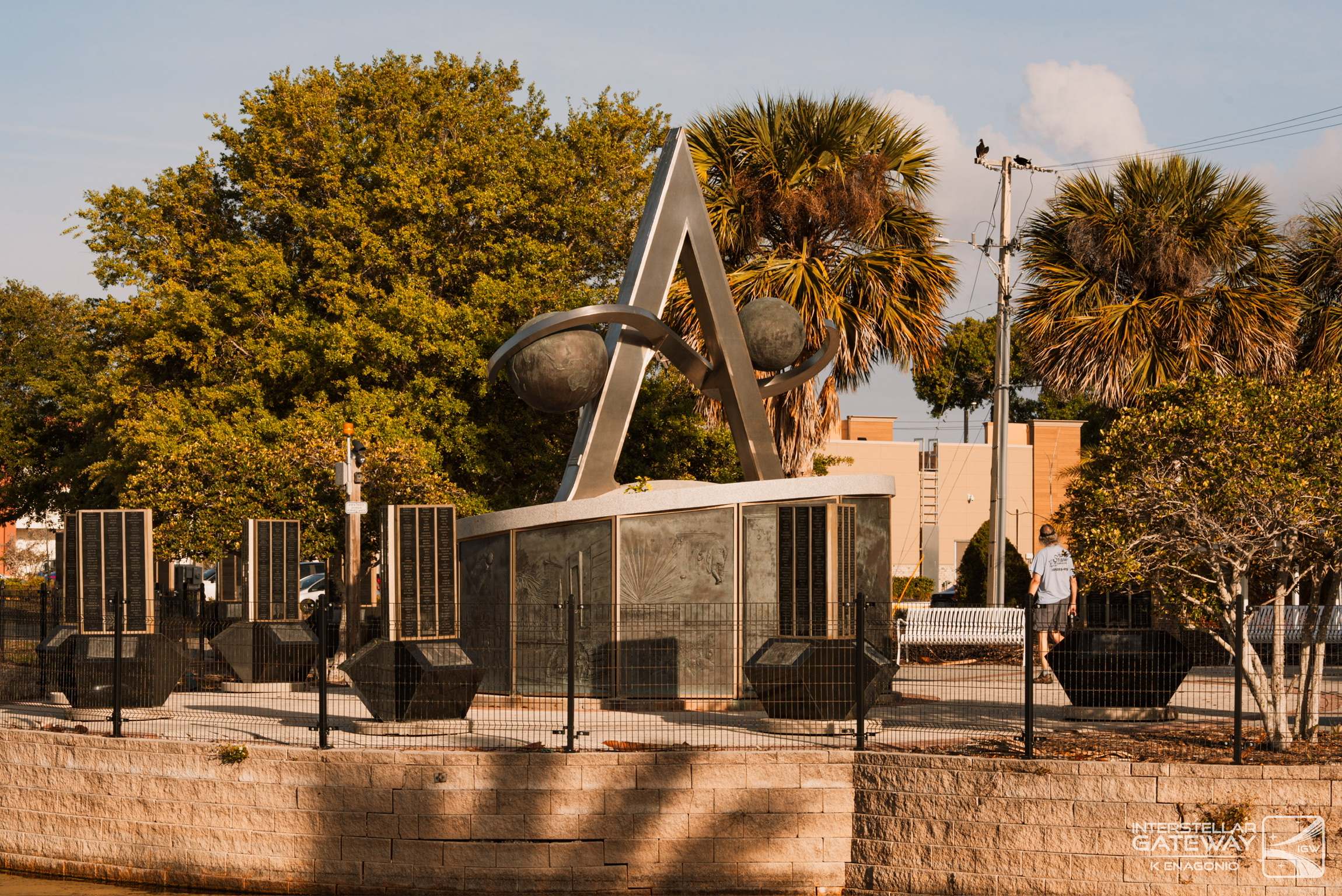 A stone monument with a stainless sculpture by the water, palms behind it.