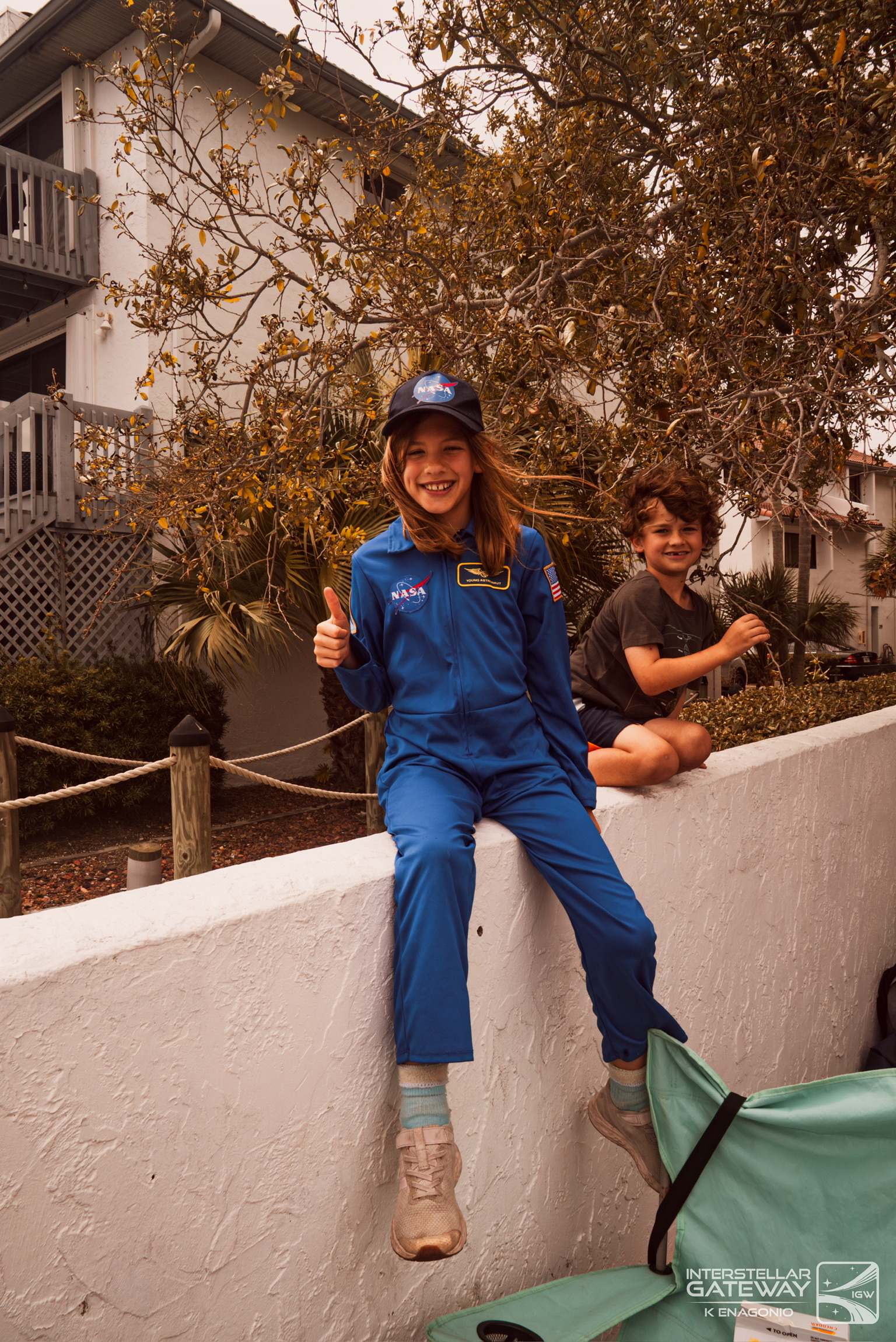 Two children, one in a blue flight suit, sit on a wall at a viewing site giving a thumbs-up.