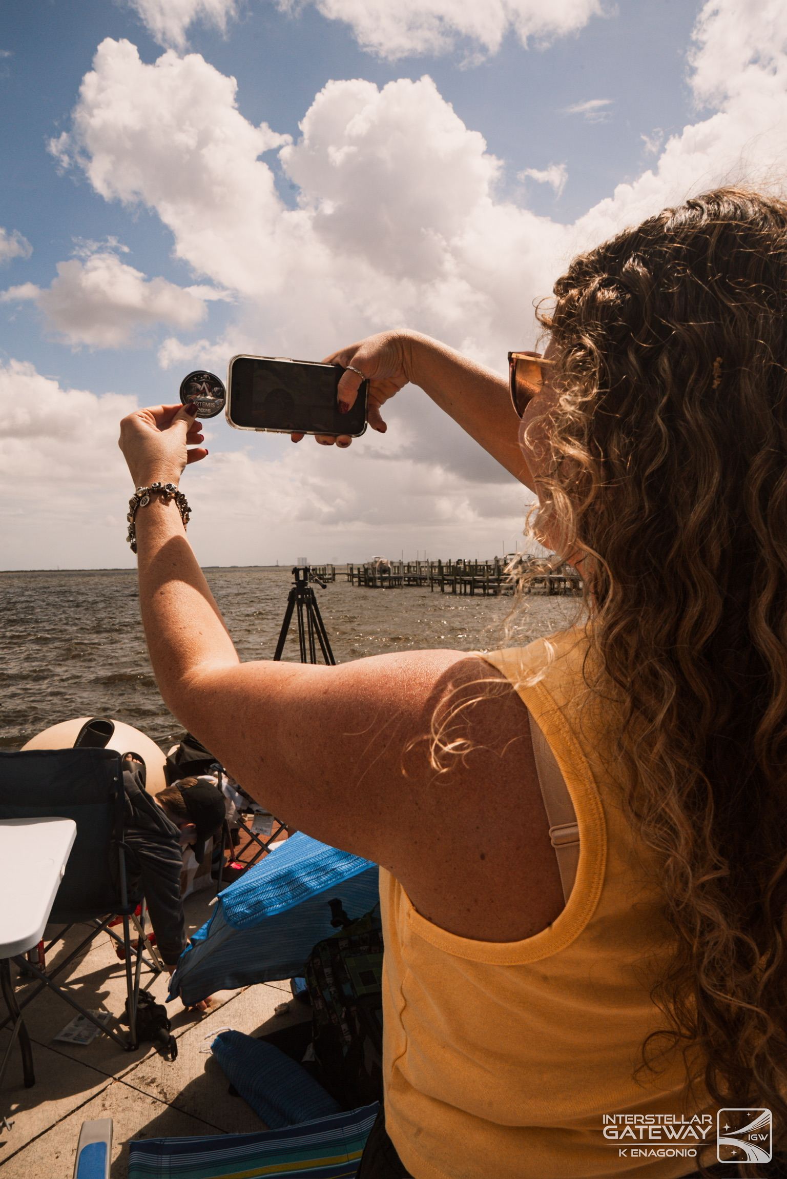 A spectator photographs the horizon with a phone from a pier under a cloudy sky.
