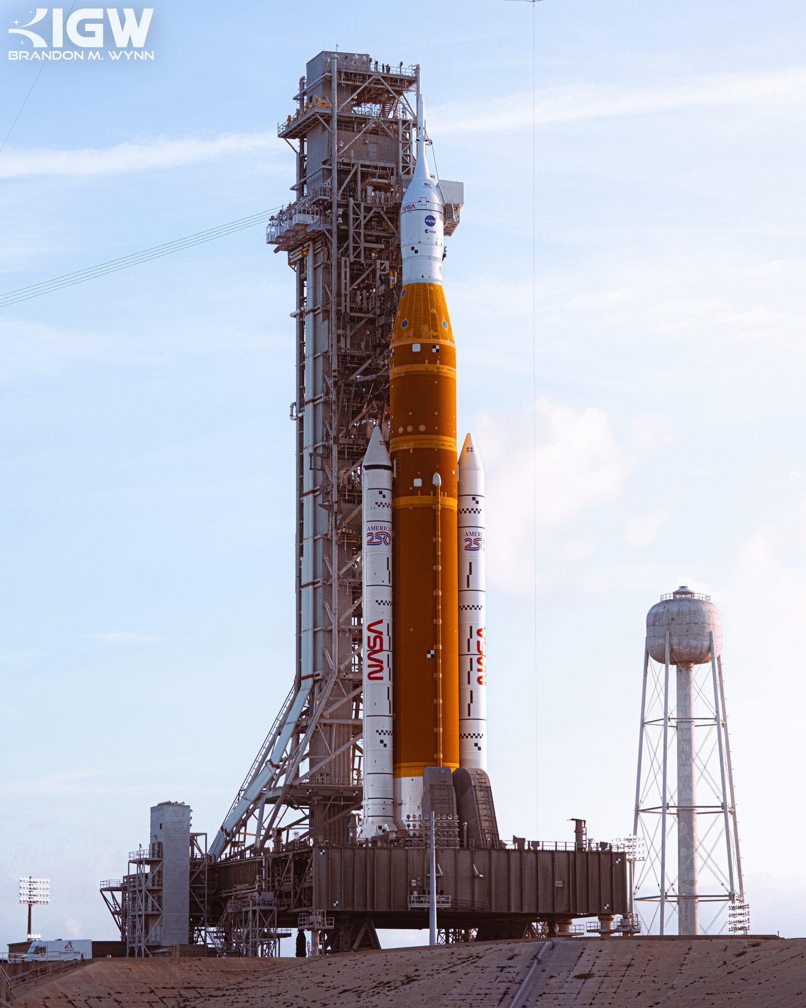 The Space Launch System on its mobile launcher in daylight, a water tower beside it.