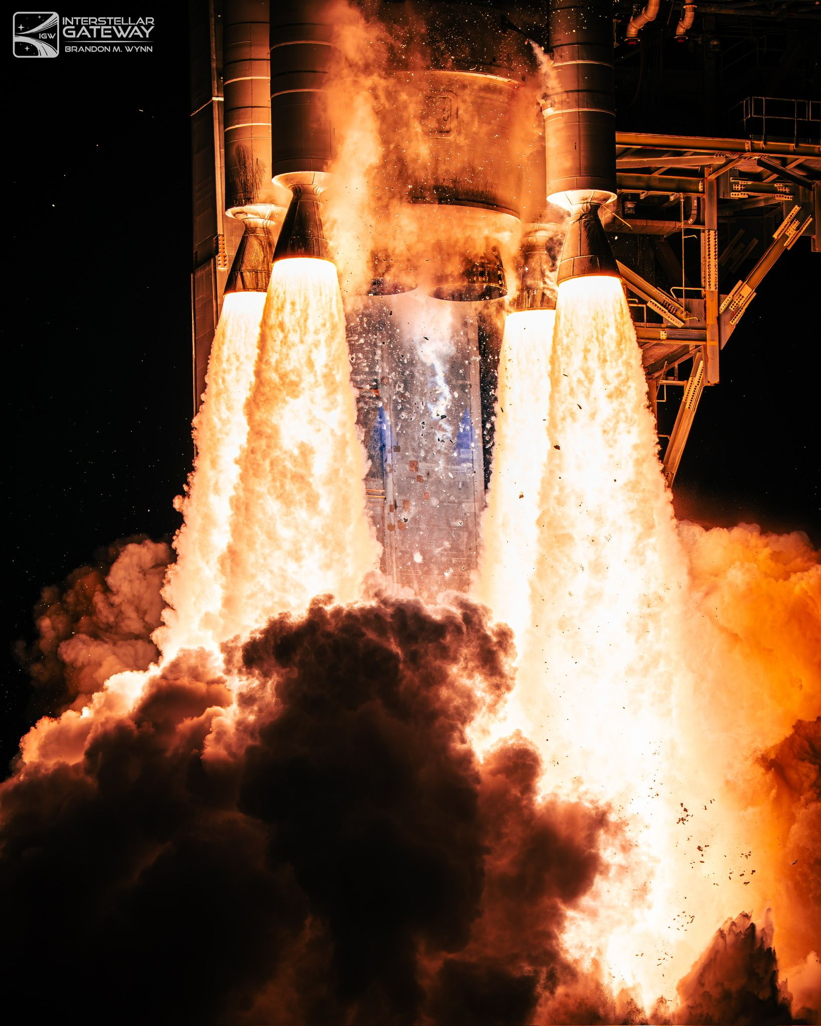A rocket lifts off at night, twin exhaust columns lighting the tower beside it.