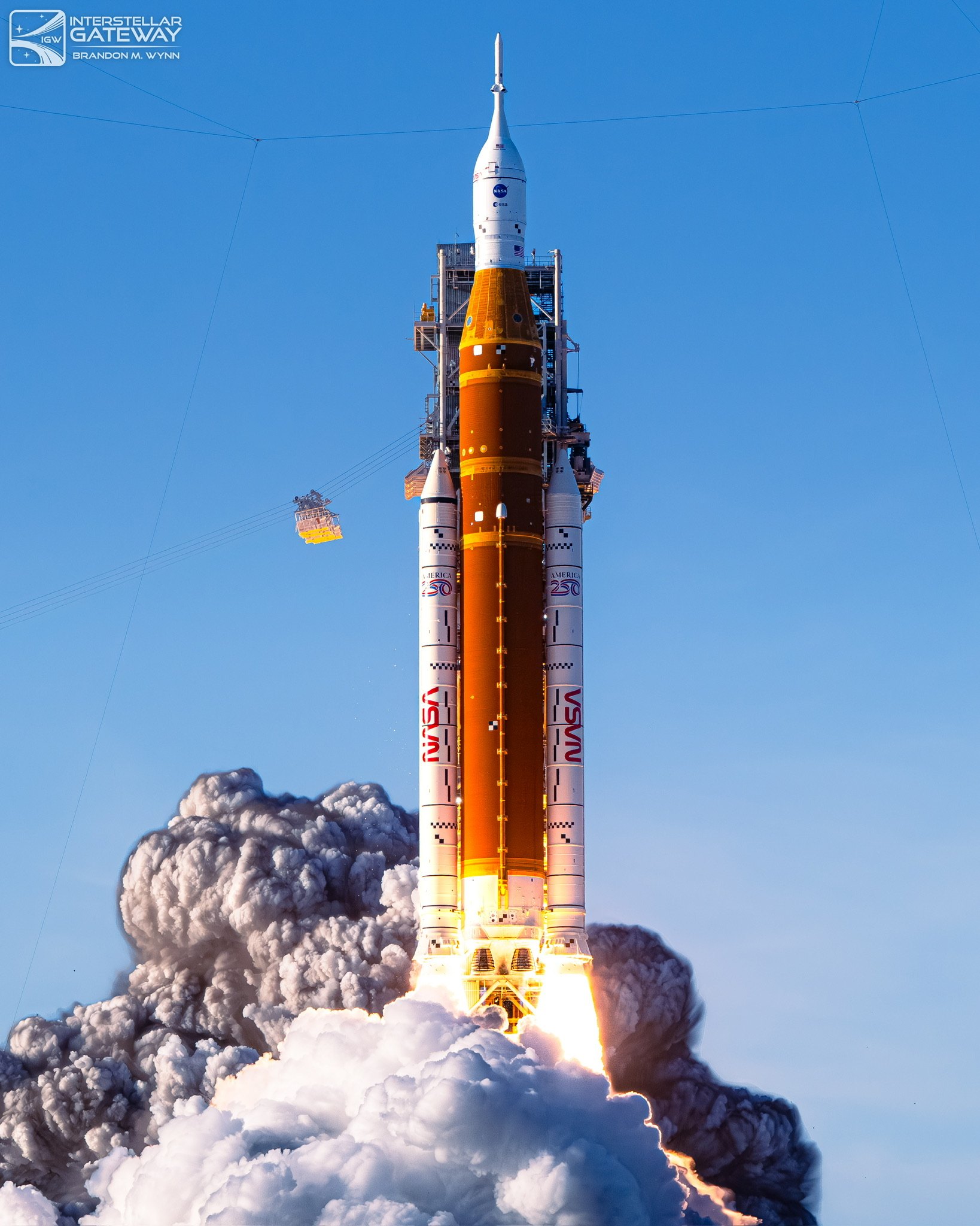 The Space Launch System climbs on a column of flame, boosters burning, the exhaust cloud spreading below.