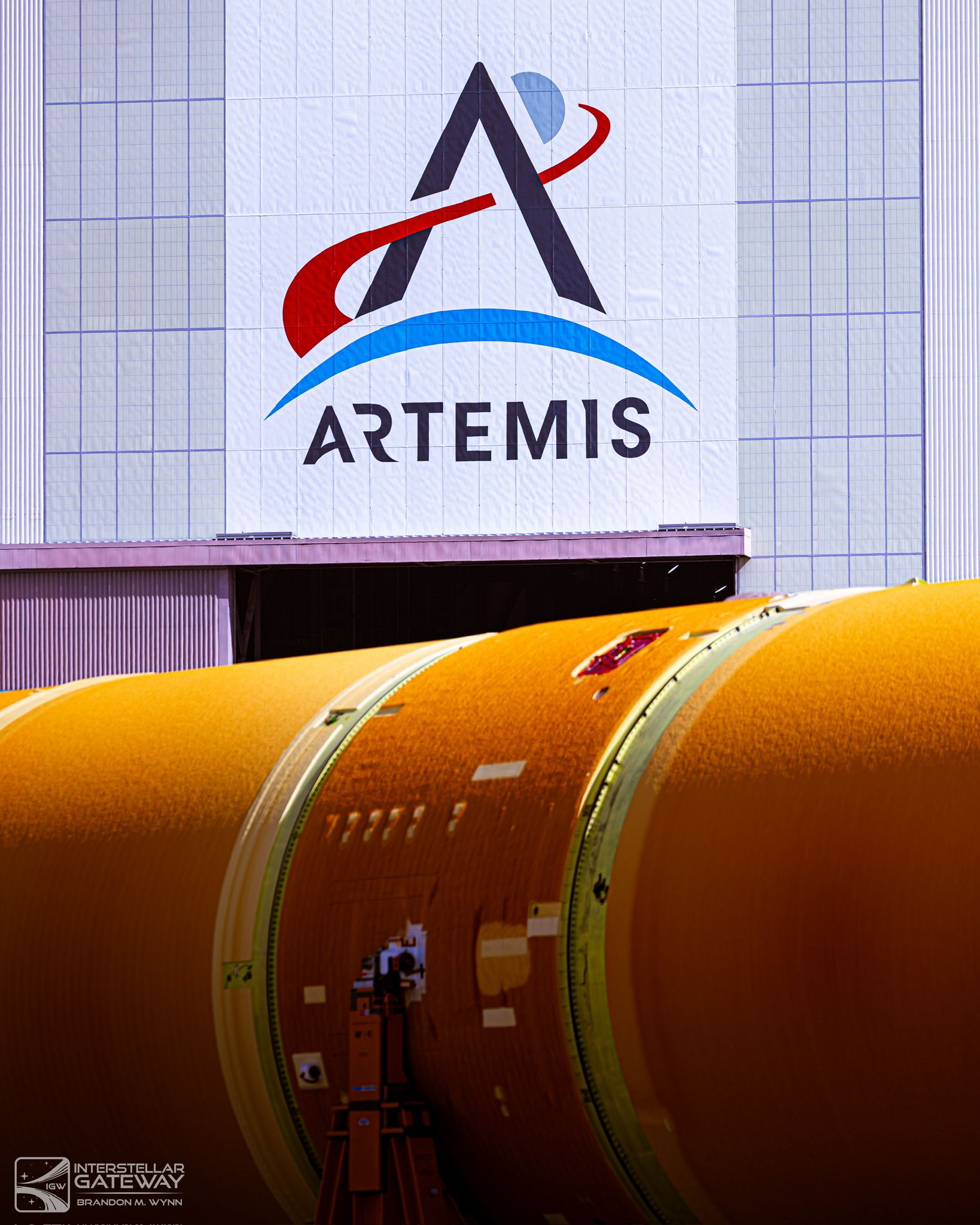 The Artemis logo on the Vehicle Assembly Building above an orange Space Launch System core stage section.