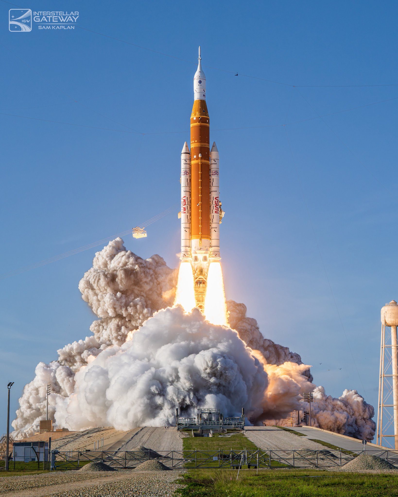 An orange core stage flanked by two white solid rocket boosters lifts off from a launch pad, driving a wall of white and grey smoke out across the ground. A water tower stands at the right edge of the frame.