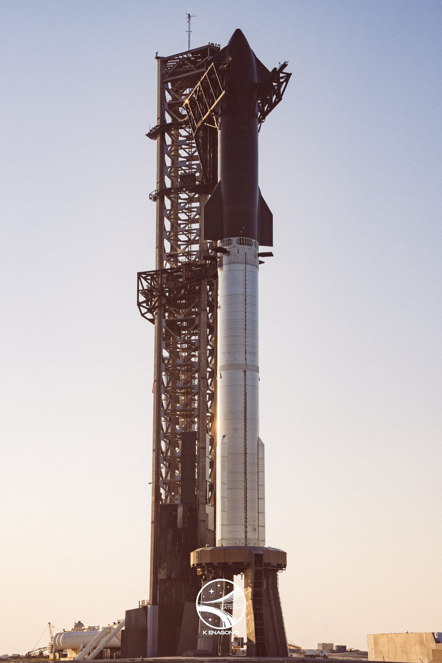 A Starship stacked on a Super Heavy booster stands beside a tall steel launch tower in low golden light, the ship's black heat-shield tiles facing the camera and the booster sitting on its launch mount.