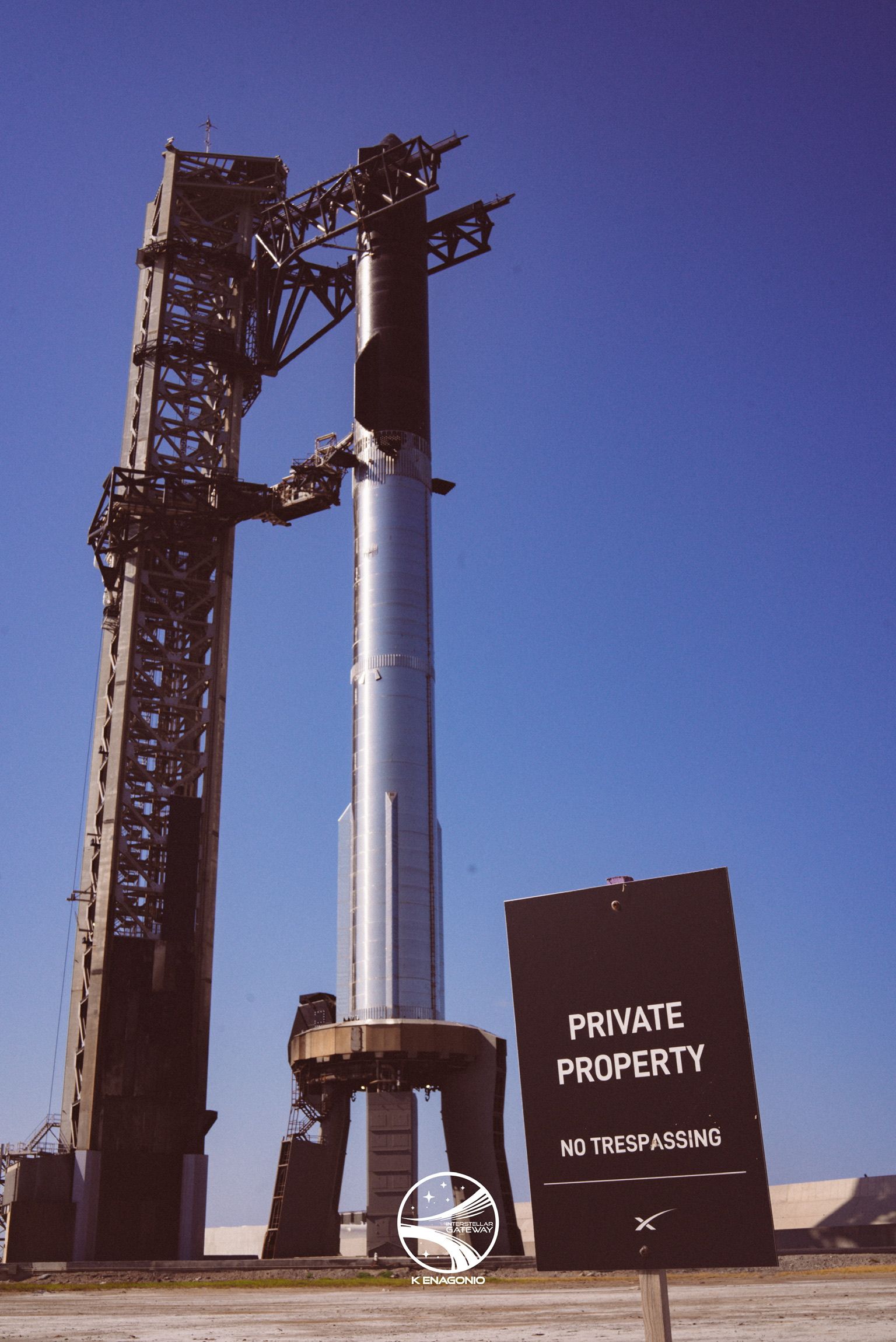 A tall stainless-steel rocket standing on its launch mount beside a steel tower fitted with catch arms, under a clear deep-blue sky. In the foreground a dark sign reads PRIVATE PROPERTY, NO TRESPASSING.