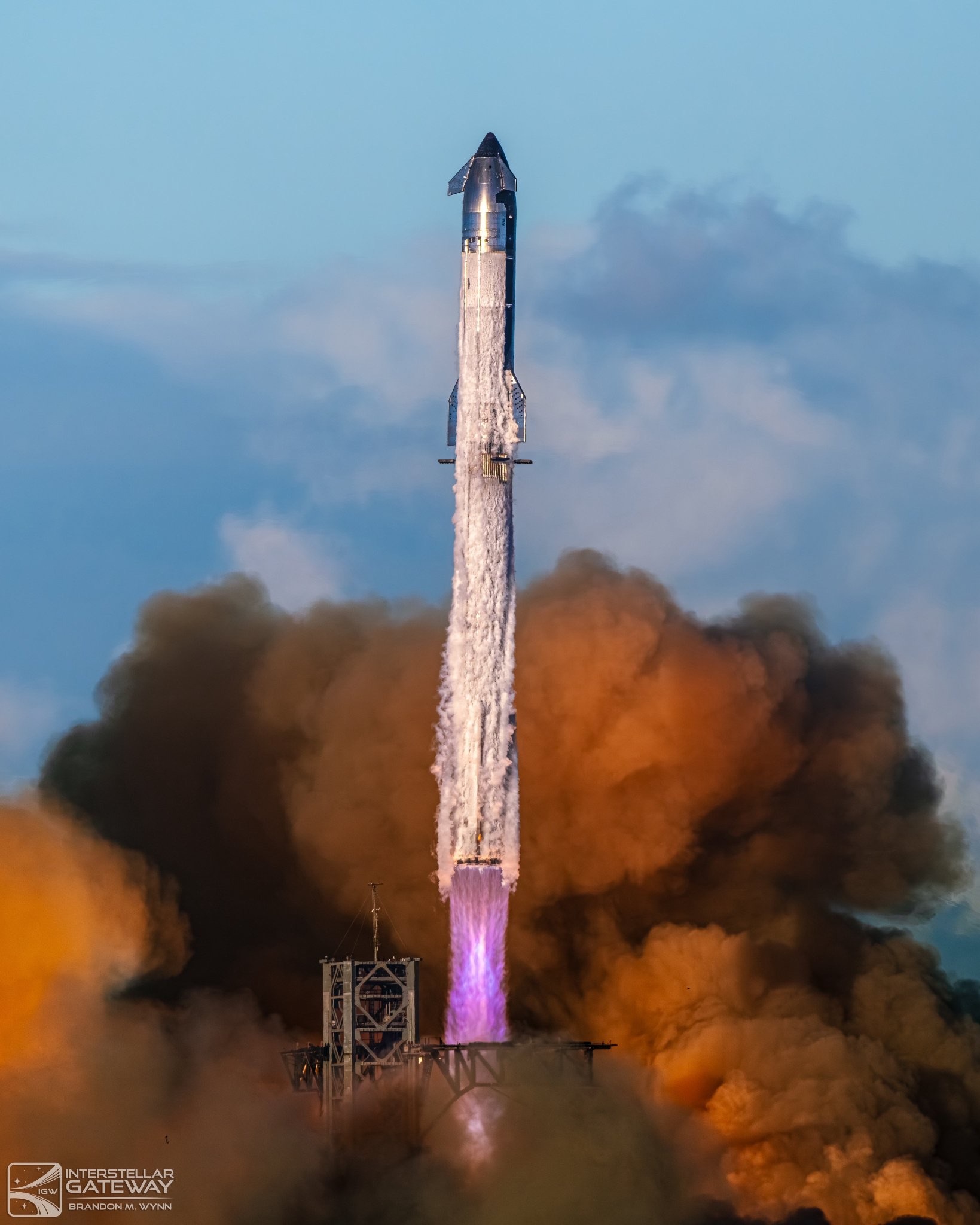 A Starship vehicle climbing away from its pad on a brilliant violet exhaust plume, condensation streaming back along its stainless flank and black-tiled nose and flaps, above huge clouds of orange- and grey-lit smoke.