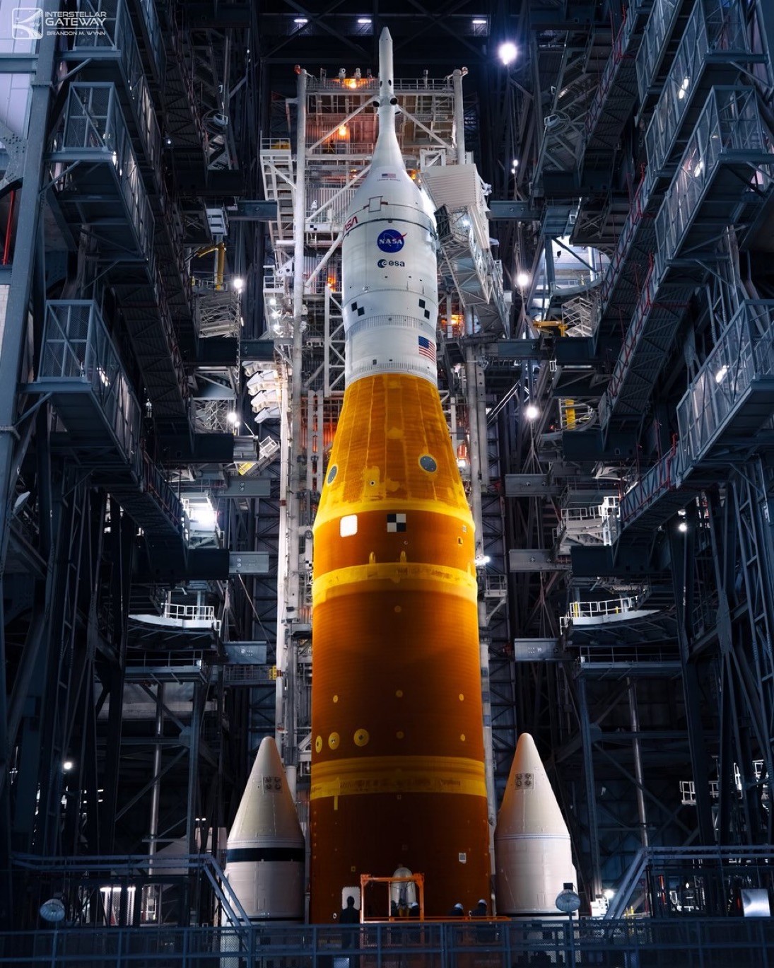 The Space Launch System standing fully stacked inside an assembly building high bay: an orange core stage flanked by two white solid rocket boosters, topped by a white crew stage carrying NASA and ESA markings and a US flag, surrounded on both sides by tiers of steel work platforms and lights.