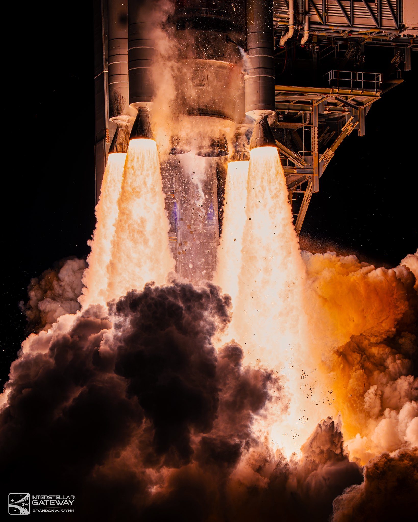The base of a rocket at ignition seen at night: four bell-shaped engine nozzles firing long white plumes, ice and debris shedding from the stage above, with the steel pad structure at right and dark and orange-lit smoke boiling up from below.