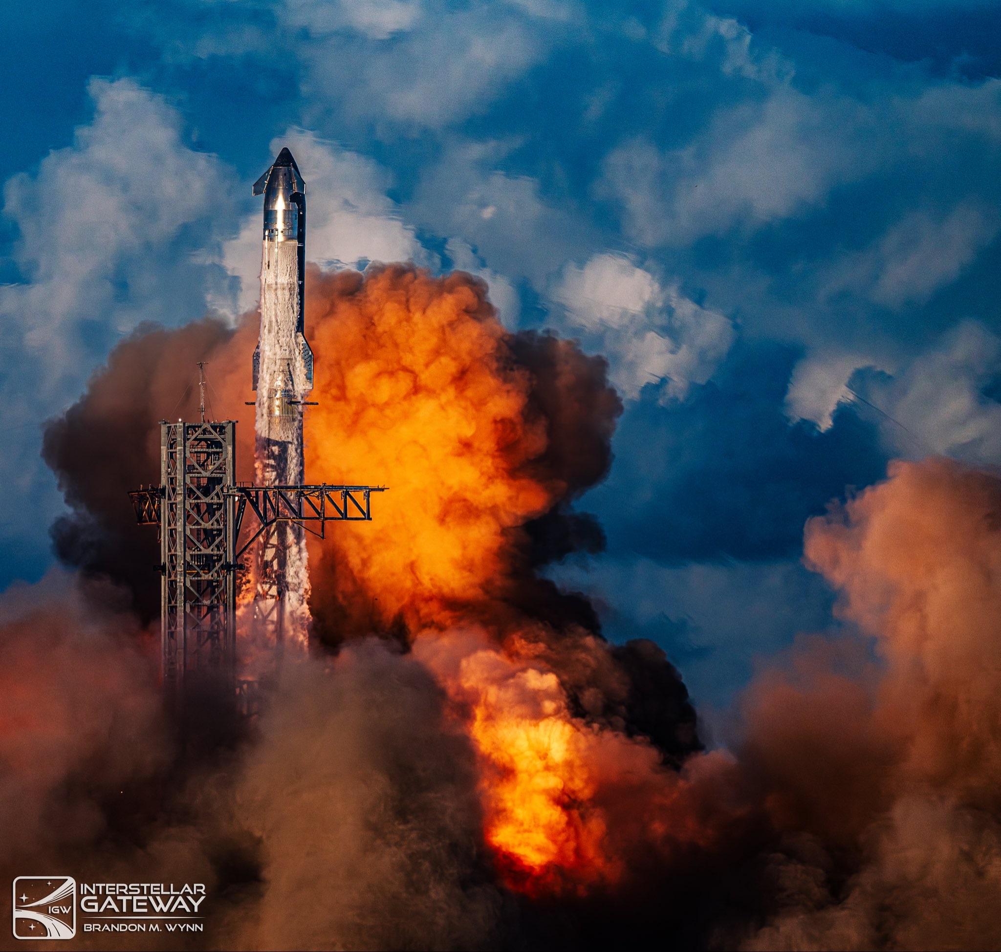 A stacked Starship and Super Heavy climbing away from the launch mount beside a lattice tower, its base wrapped in a vast orange fireball and dark billowing exhaust under a blue, cloud-streaked sky.