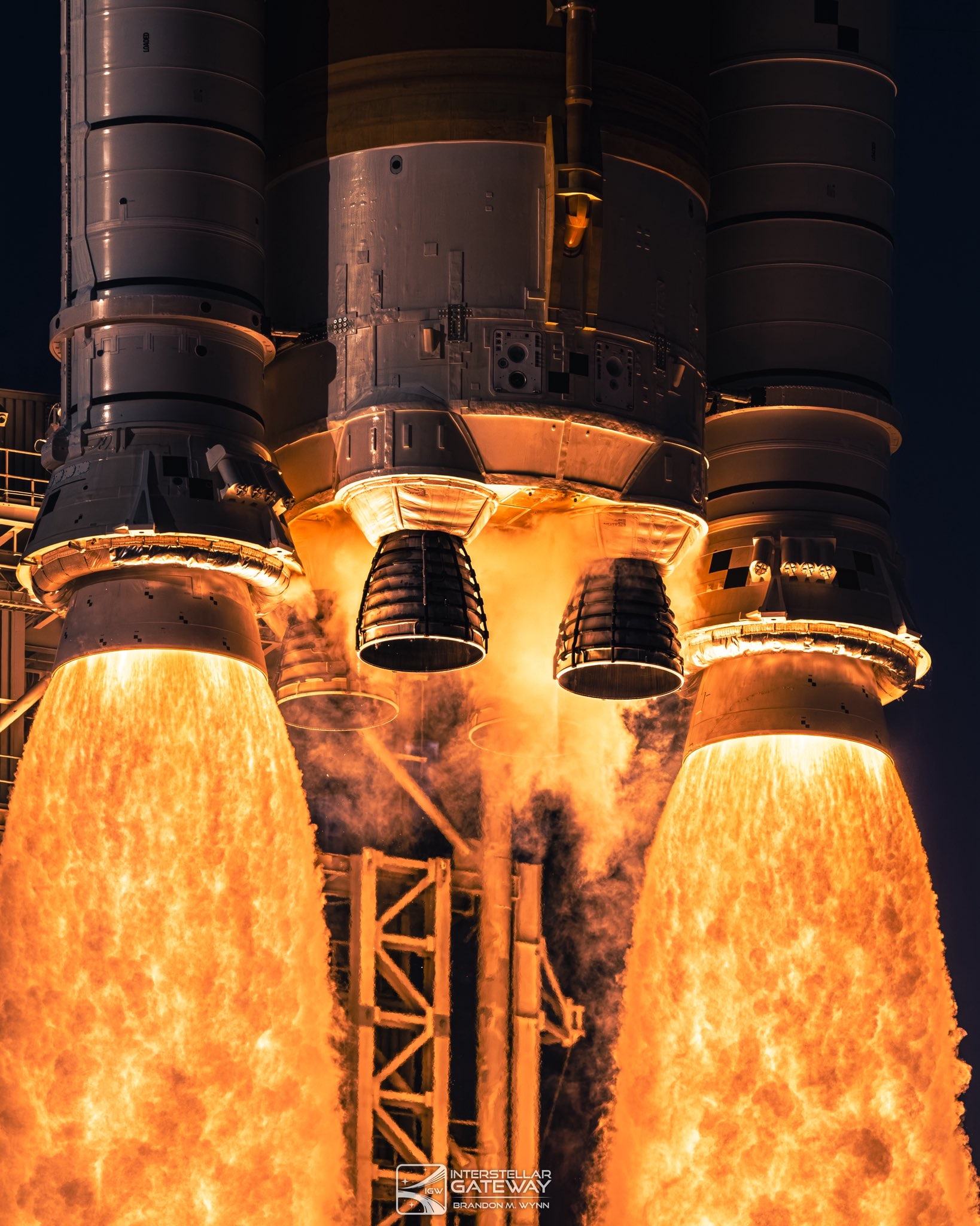 Close view of a rocket's engine section at liftoff: bell nozzles clustered under the core stage between two solid rocket boosters, all pouring brilliant orange flame past the launch structure into a dark sky.