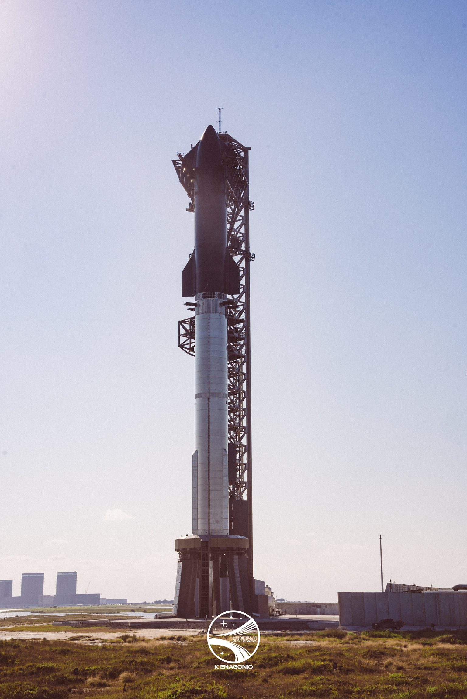 A fully stacked Starship and Super Heavy standing on its launch mount beside the lattice tower, seen across flat scrub and sandy ground, with shallow water and wetland flats beyond and tall pale buildings on the distant horizon.