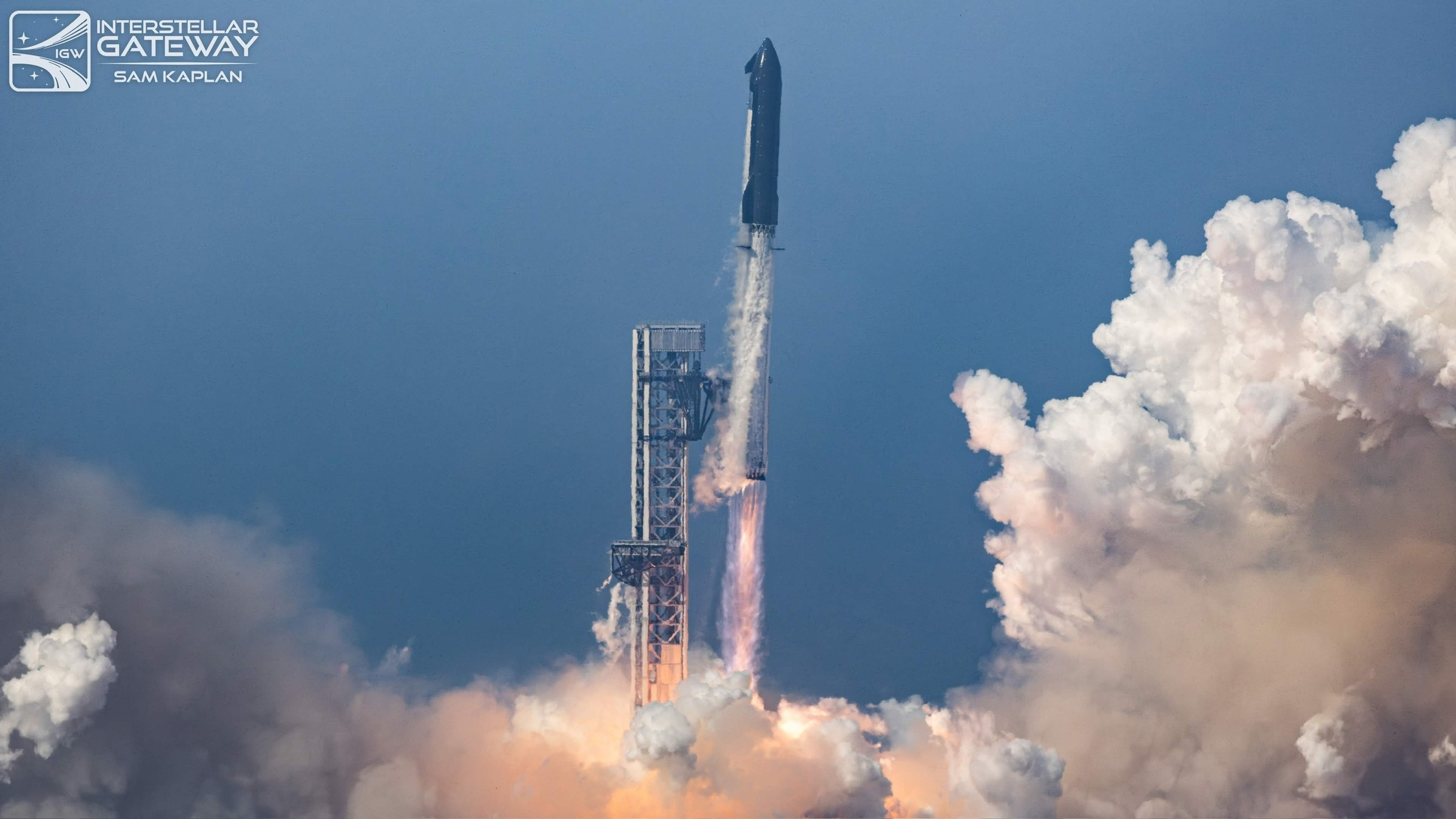 A stacked Starship and Super Heavy rising just past the top of its lattice launch tower in daylight, engines burning behind a rolling bank of white exhaust against a deep blue sky.