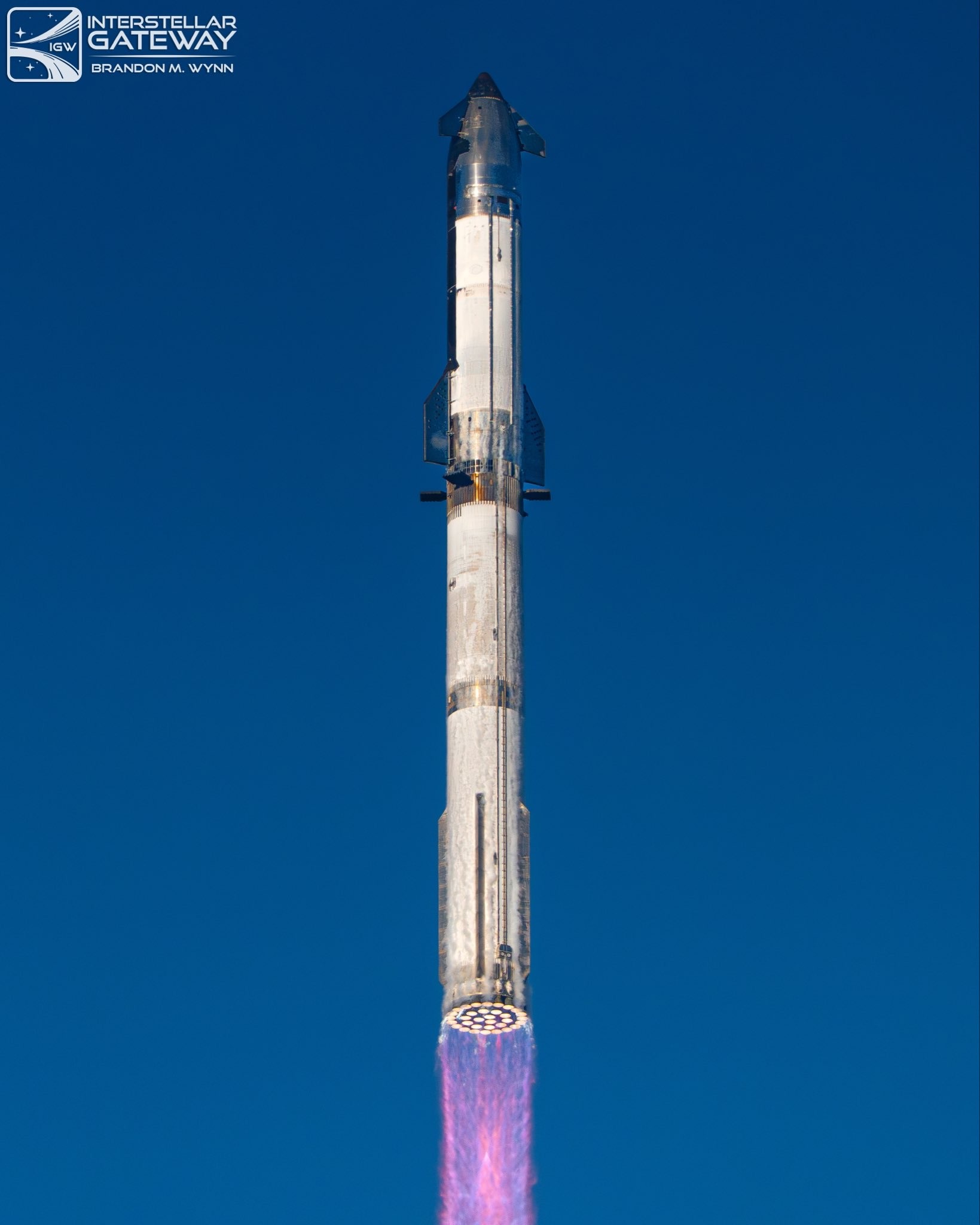 A stacked Starship and Super Heavy in powered flight against a deep blue sky, seen head-on with the ship's forward and aft flaps in silhouette and a violet plume streaming from the ring of lit engines at the base.