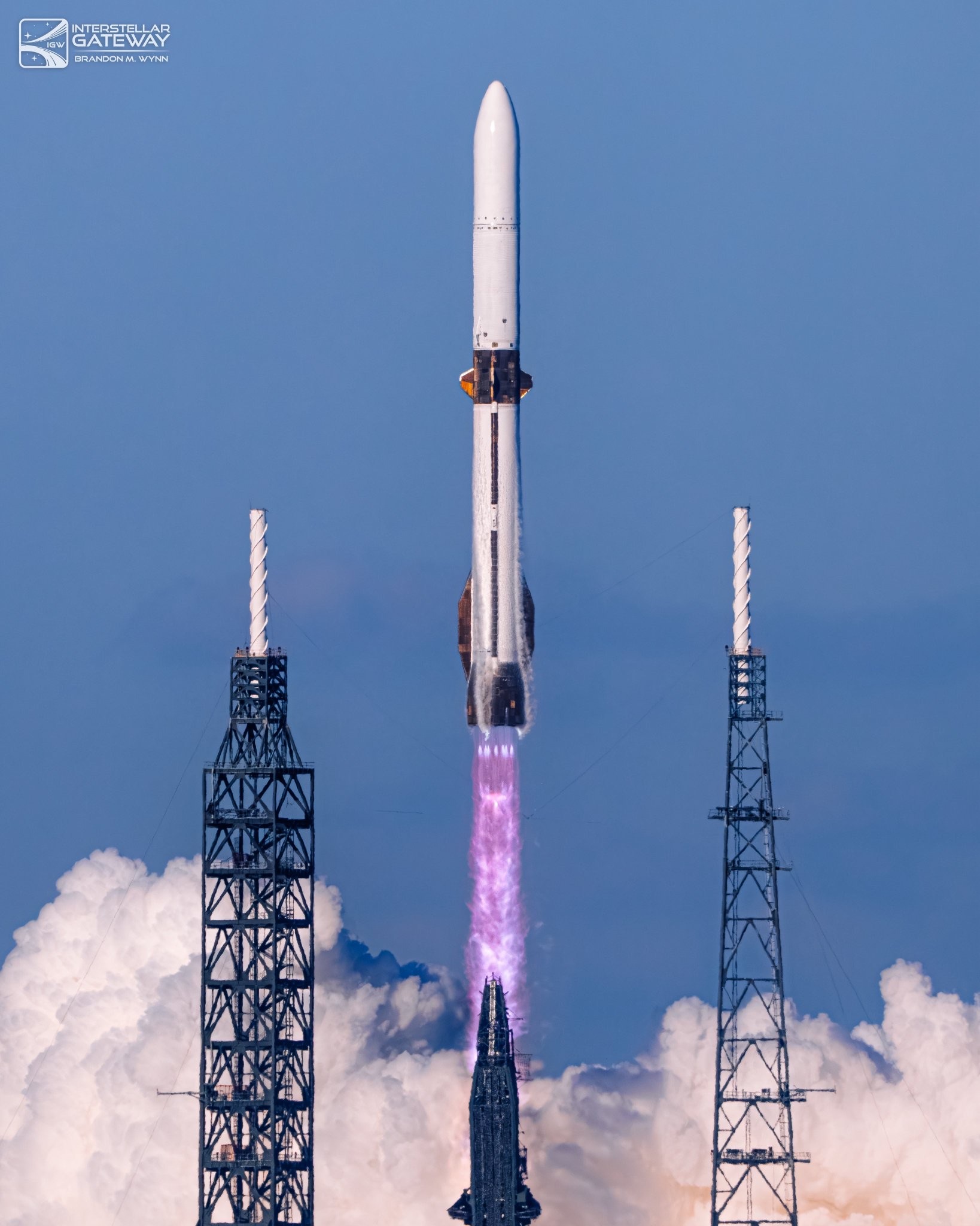 A white rocket with a white payload fairing climbs between two tall lattice lightning towers, a violet exhaust plume beneath it and sunlit smoke banked low across the base of the frame.