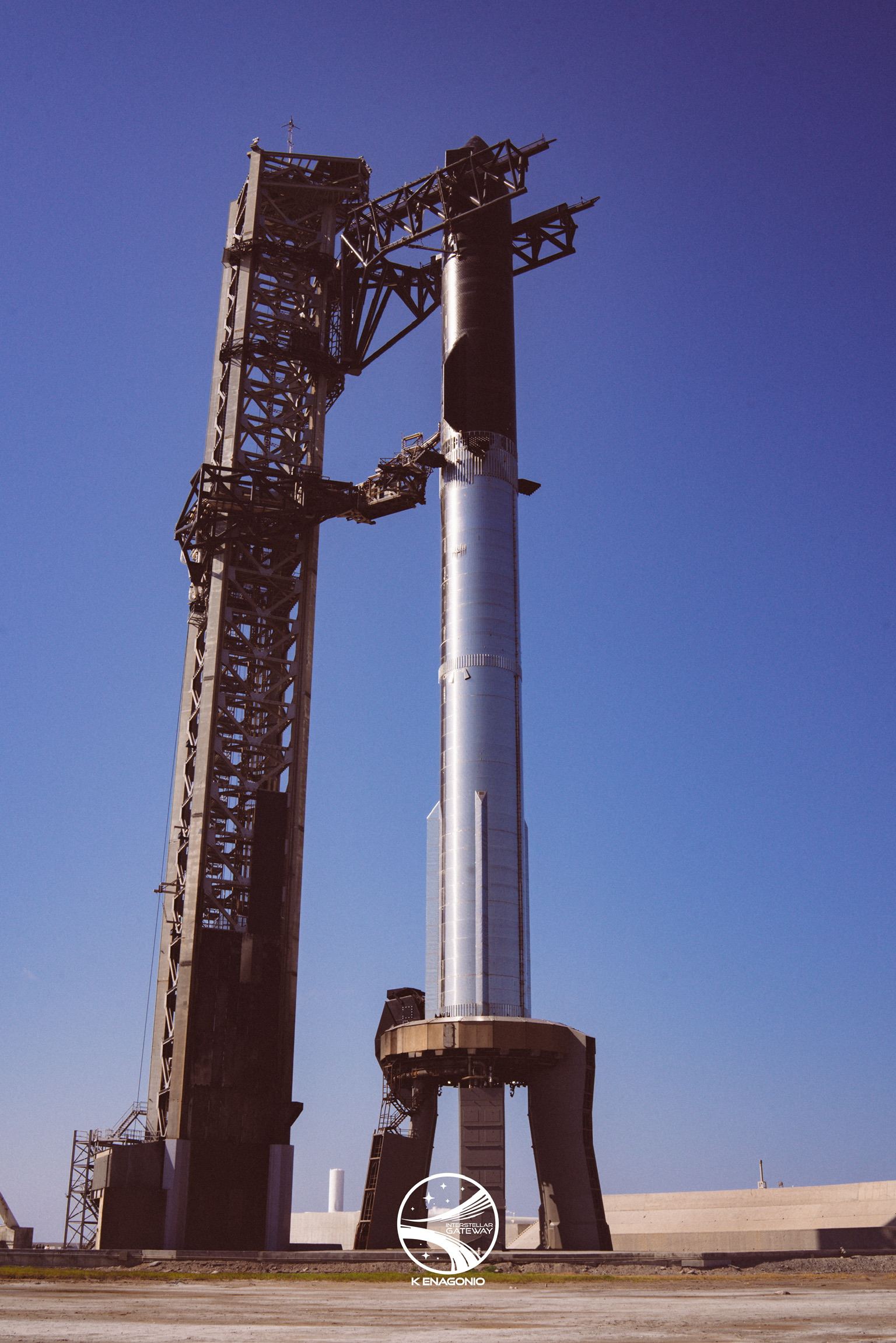 A tall stainless-steel Starship vehicle stands on a legged launch mount beside a lattice launch tower with its lifting arms raised, under a clear blue sky. The lower half of the vehicle is bare bright metal and the upper section is dark against the sky.