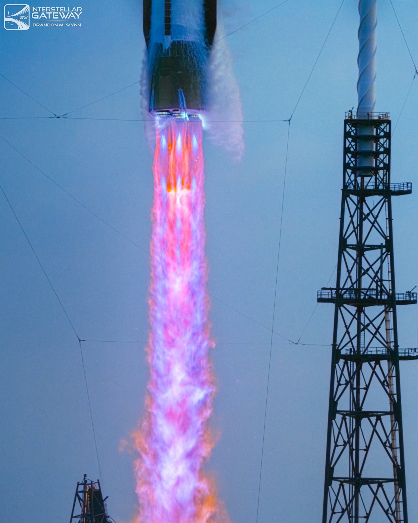 A tight crop of a rocket's engine section in early ascent, with a long pink and violet exhaust plume streaming down the frame. A lattice lightning tower and taut guy wires stand against the pale twilight sky at right.