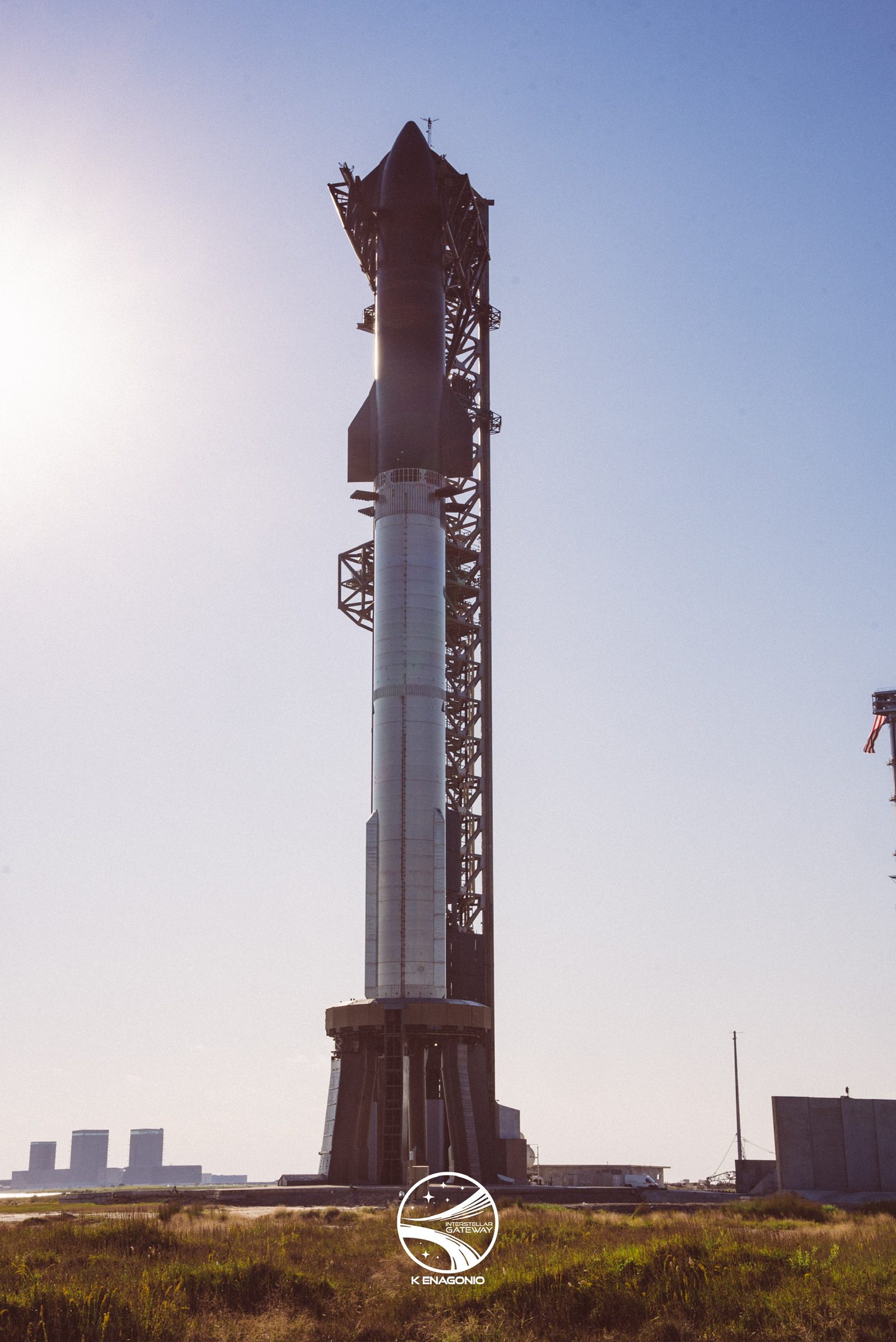A fully stacked Starship and Super Heavy standing on its launch mount, the dark upper stage and its forward flaps silhouetted against a hazy sky with the sun flaring at the left edge. The launch tower's steelwork rises alongside, and flat grassland runs out to storage tanks on the distant horizon.