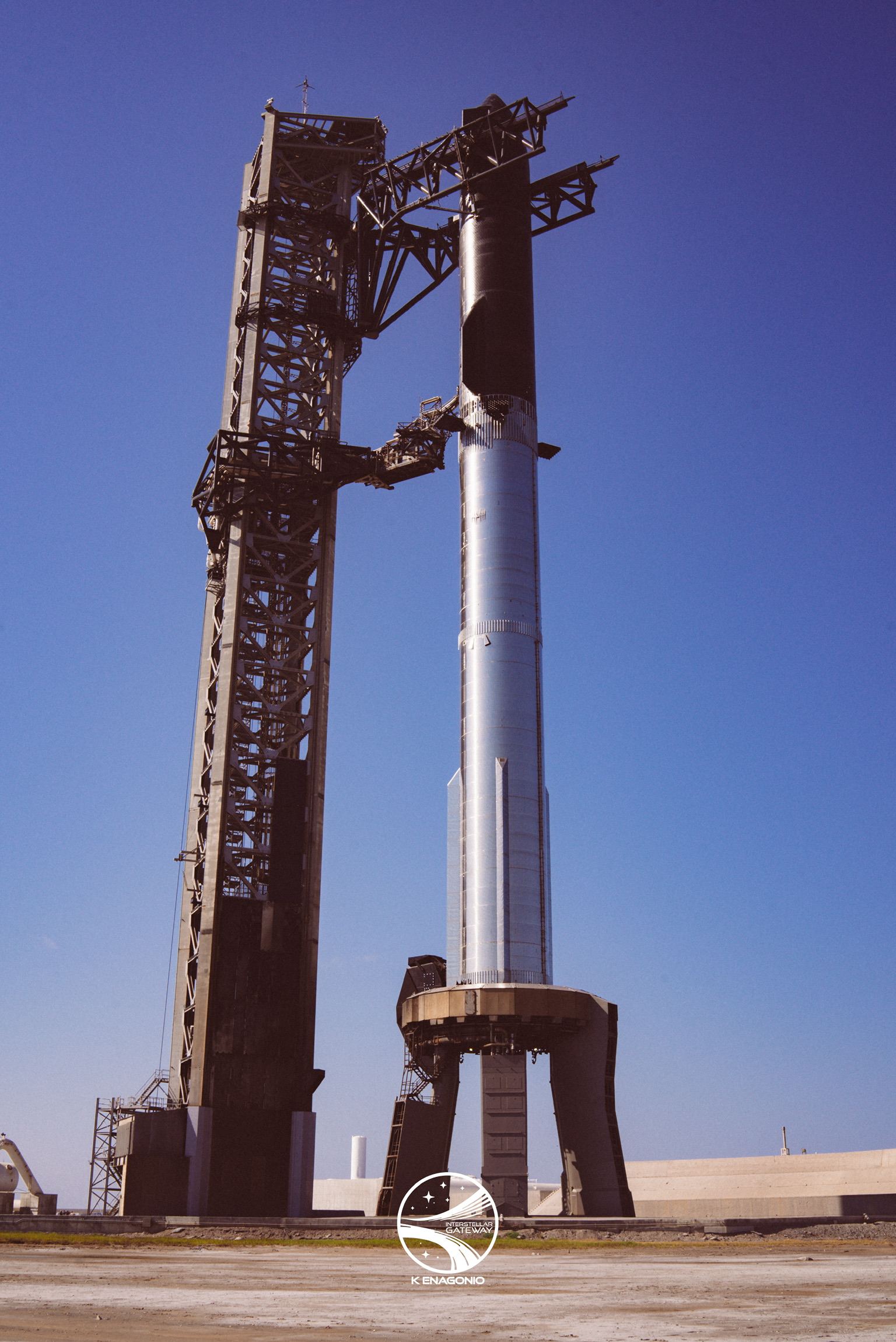 A stainless-steel Super Heavy booster standing on its launch mount with a dark, heat-shield-tiled Starship above it, seen from ground level under a clear blue sky. The launch tower rises at left with its two arms reaching across toward the vehicle.