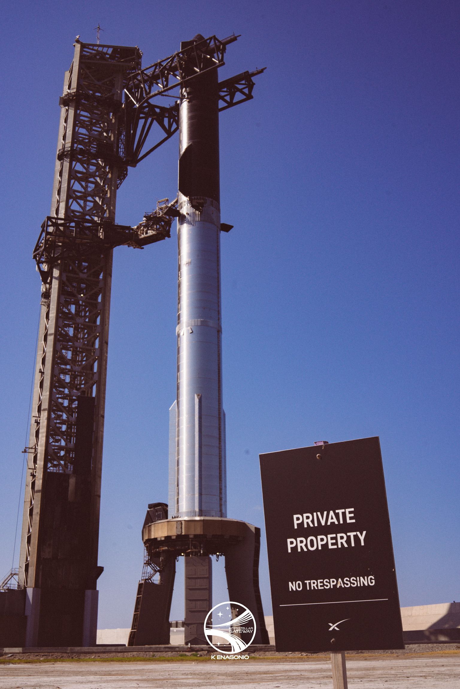 A stacked Starship and Super Heavy standing beside its launch tower under a clear blue sky, with a dark sign in the right foreground reading PRIVATE PROPERTY, NO TRESPASSING.