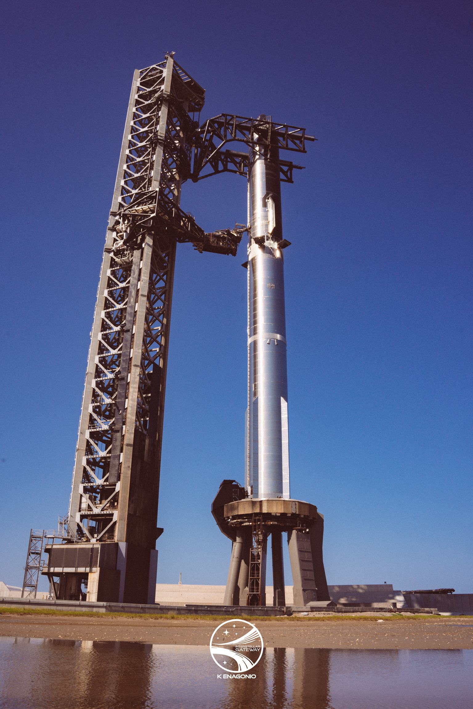 A bare stainless-steel Super Heavy booster standing alone on its launch mount beside the launch tower under a deep blue sky, mirrored in shallow water across the foreground.