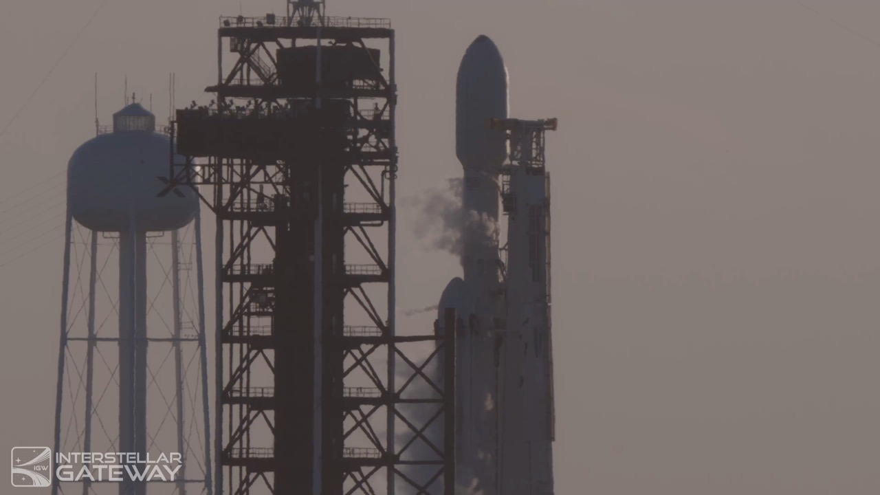 A rocket standing at its pad in flat hazy light, a payload fairing at the top and a second rounded nose cone lower down and offset, with vapour venting from its side, a tall lattice service structure alongside it and a water tower at the left of the frame.