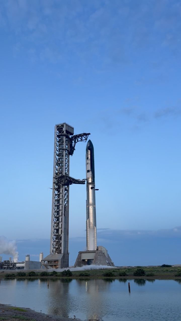 A stacked Starship and Super Heavy stand on the launch mount beside a tall steel launch tower, venting white vapour at the base, with the pair reflected in a flat stretch of water in the foreground under a pale blue sky.