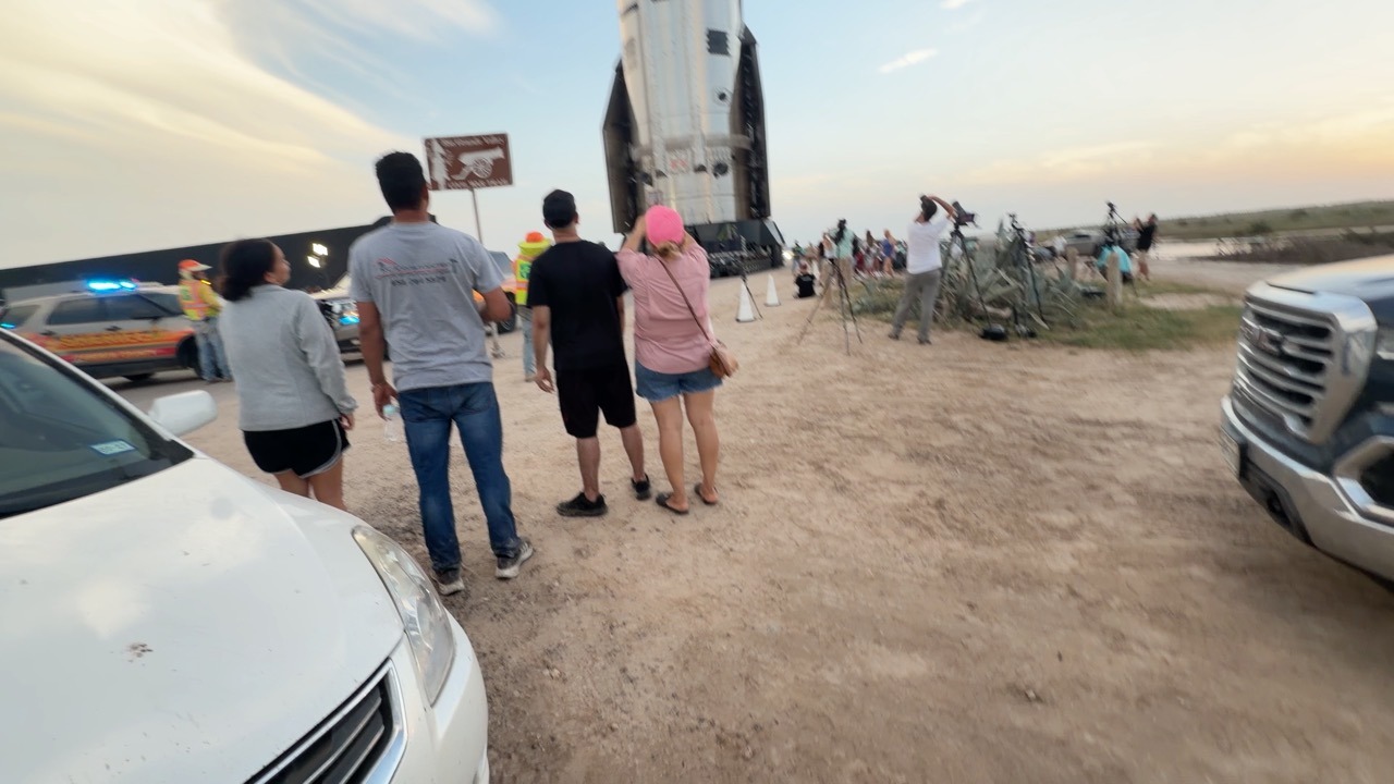 A group of onlookers stands on a dirt roadside beside parked cars and a vehicle with flashing lights, watching a stainless-steel rocket stage on a wheeled transporter, while photographers work from tripods further along the verge under a low sun.