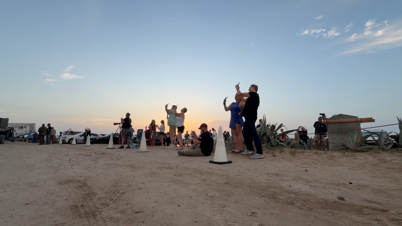 Spectators spread across a sandy lot with the sun low on the horizon, several holding phones up to film, with traffic cones, parked cars, camera tripods and agave plants around them under a wide orange and blue sky.