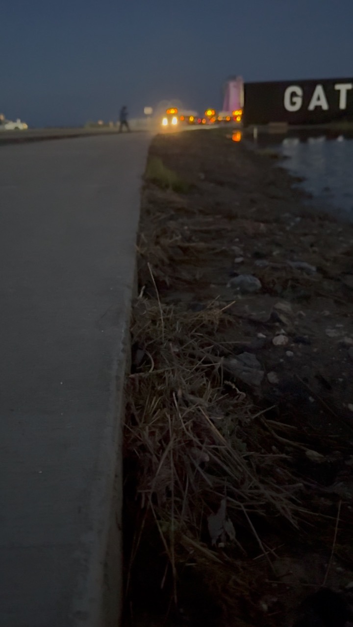 A low view along the edge of a paved road at dusk, dry grass and stones sloping down to water on the right. Vehicle lights and a distant walking figure are visible up the road, with a large dark sign carrying white lettering beyond them.