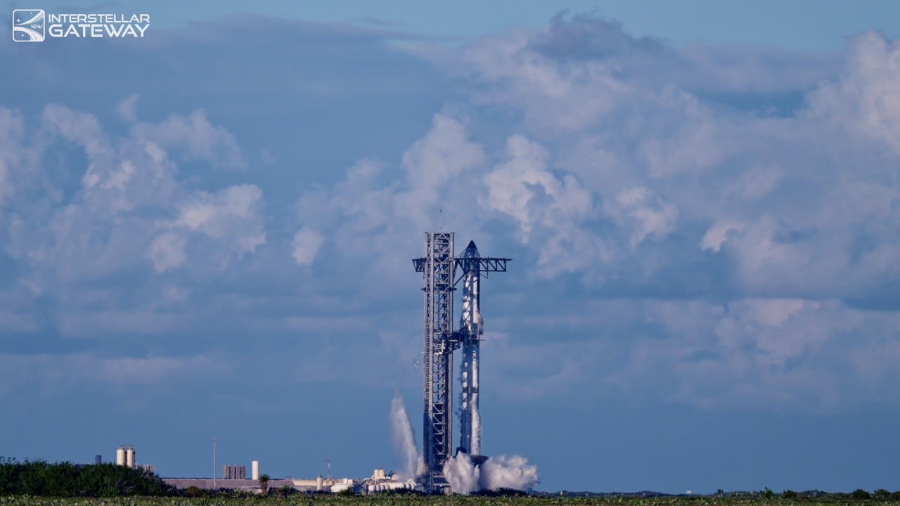 A stacked Starship and Super Heavy stand beside their launch tower under a blue sky filled with tall white cloud, with white vapour venting around the base of the vehicle.