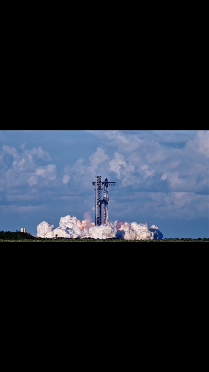 A stacked Starship and Super Heavy fire their engines beside the launch tower in daylight, flame at the base and a wide bank of white exhaust rolling out across flat ground under a partly cloudy blue sky.