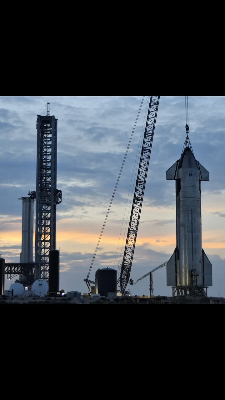 A stainless Starship upper stage with its nose cone and forward and aft flaps hangs from crane cables at dusk above a stand, with a tall steel lattice tower, another stainless stage, storage tanks and ground equipment behind it against an orange and blue sky.