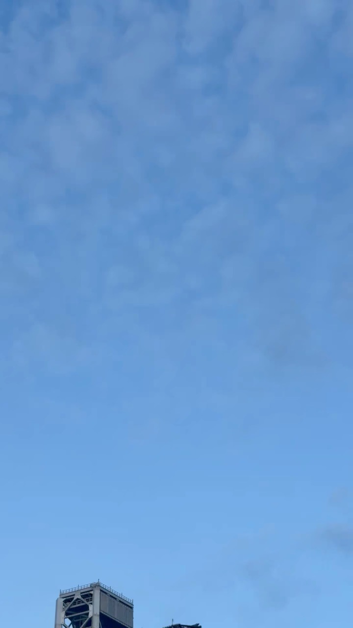 A wide expanse of pale blue sky scattered with thin cloud, with the top of a steel lattice tower just entering the very bottom of the frame.