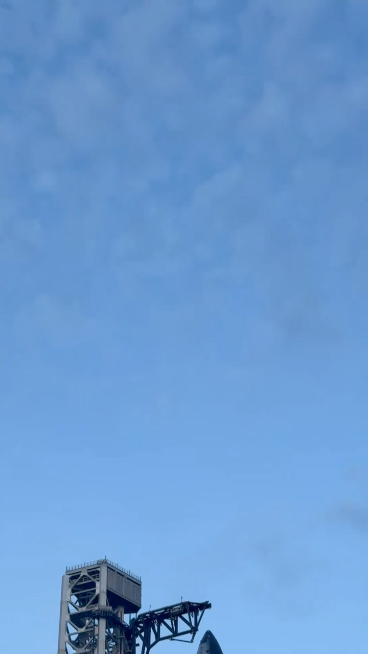 A steel launch tower with a boxed platform and a lattice service arm rises at the lower left edge of the frame, beside the dark pointed tip of a rocket. The rest of the frame is open blue sky with thin high cloud.