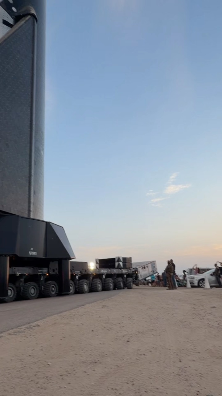 The lower section of a very large rocket, its skin covered in dark tiles, sits on a multi-axle wheeled transporter on a sandy road. A line of support trailers, vehicles and a few standing figures stretches away toward a low sun on the horizon.