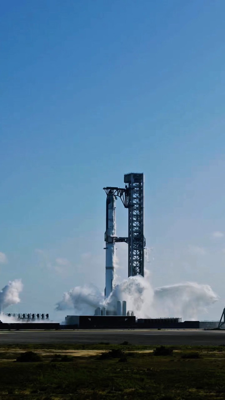 A stacked Starship and Super Heavy stands on its launch mount beside the tall lattice launch tower, white vapour billowing across the pad below it. Flat scrub and grassland fill the foreground under a clear blue sky.